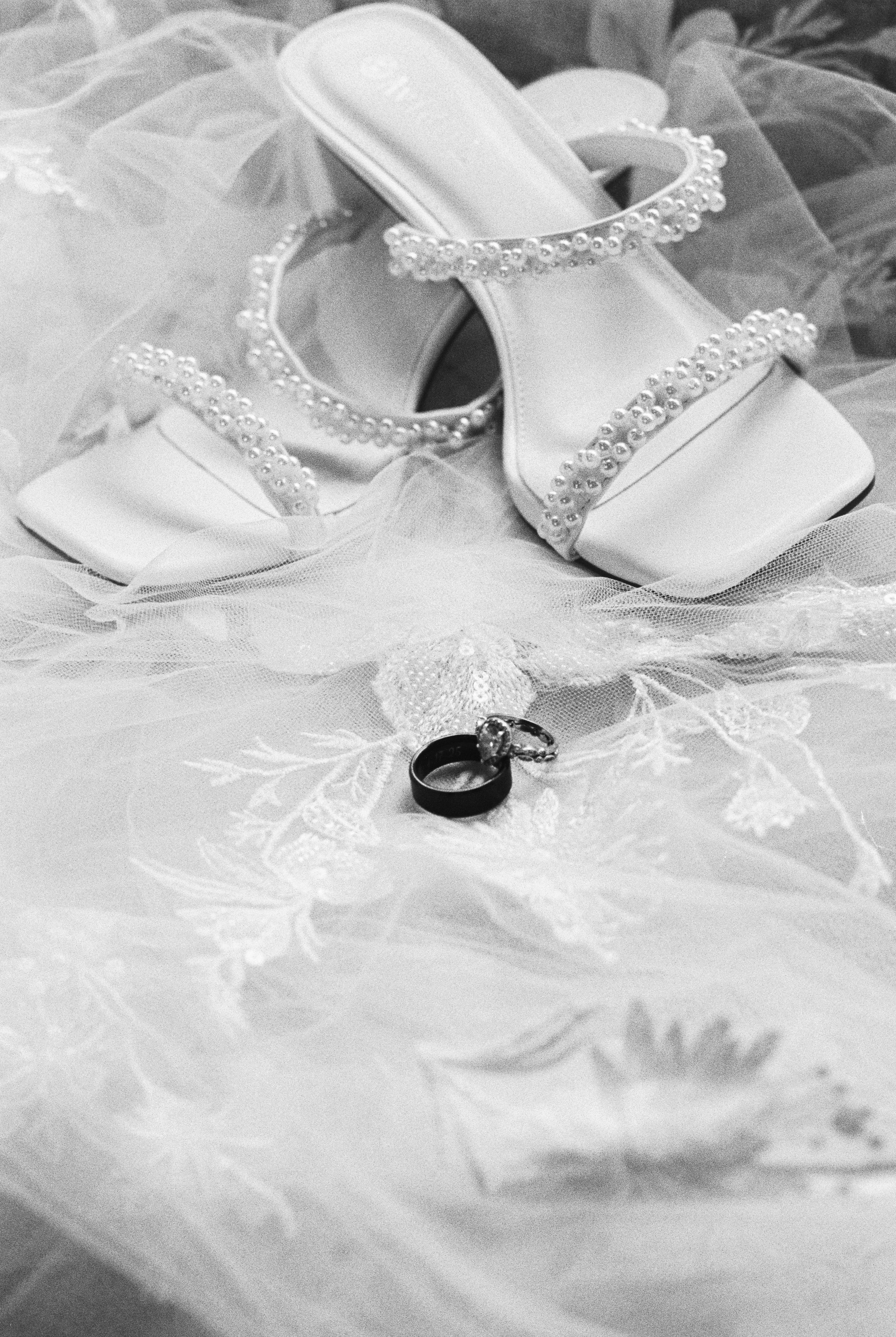 Black and white photo of a pair of wedding shoes decorated with pearls, placed on a lace fabric, with two wedding rings in front.