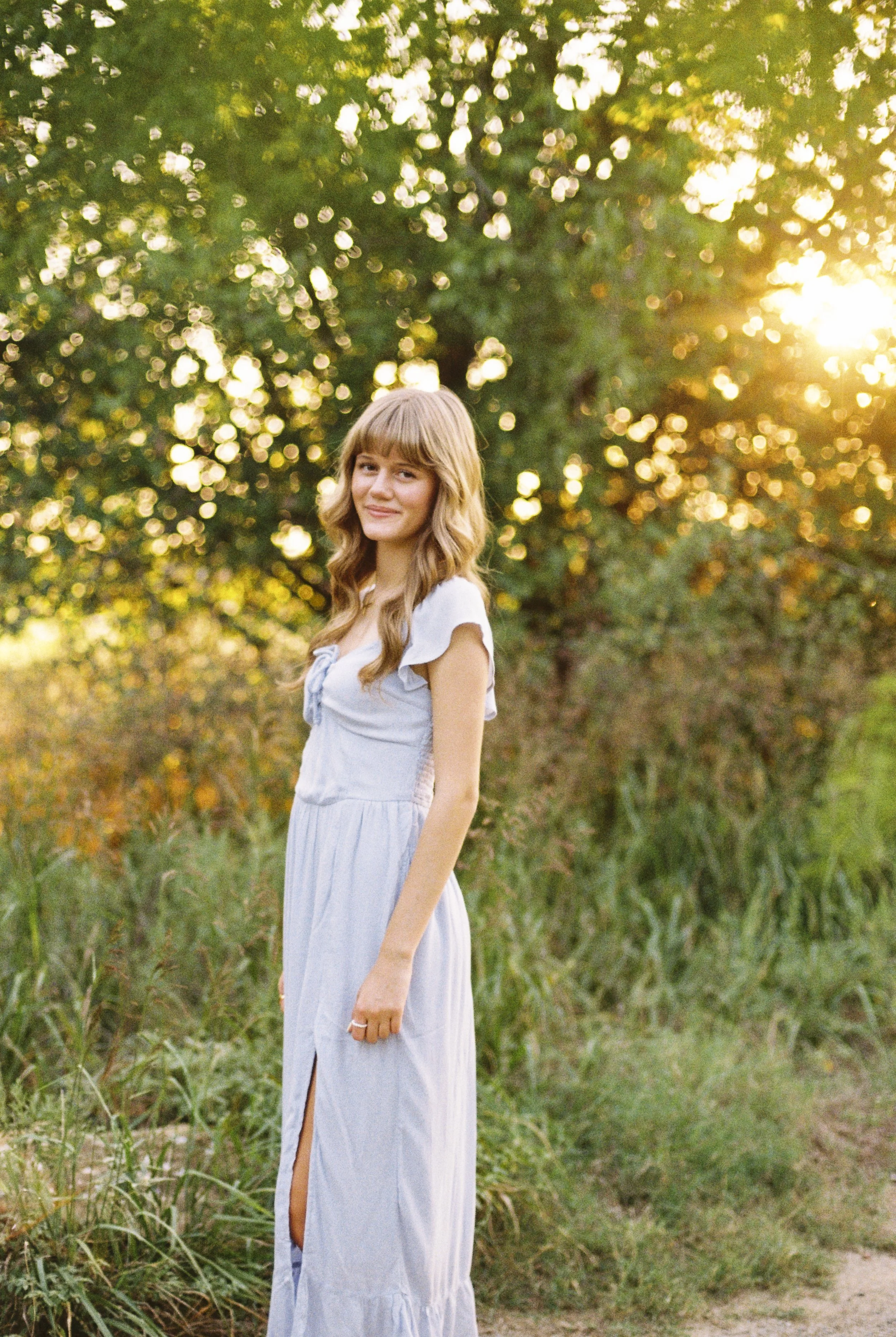 A young woman with long, wavy blonde hair wearing a light blue sleeveless maxi dress stands outdoors among green grass and trees during sunset, smiling softly at the camera.
