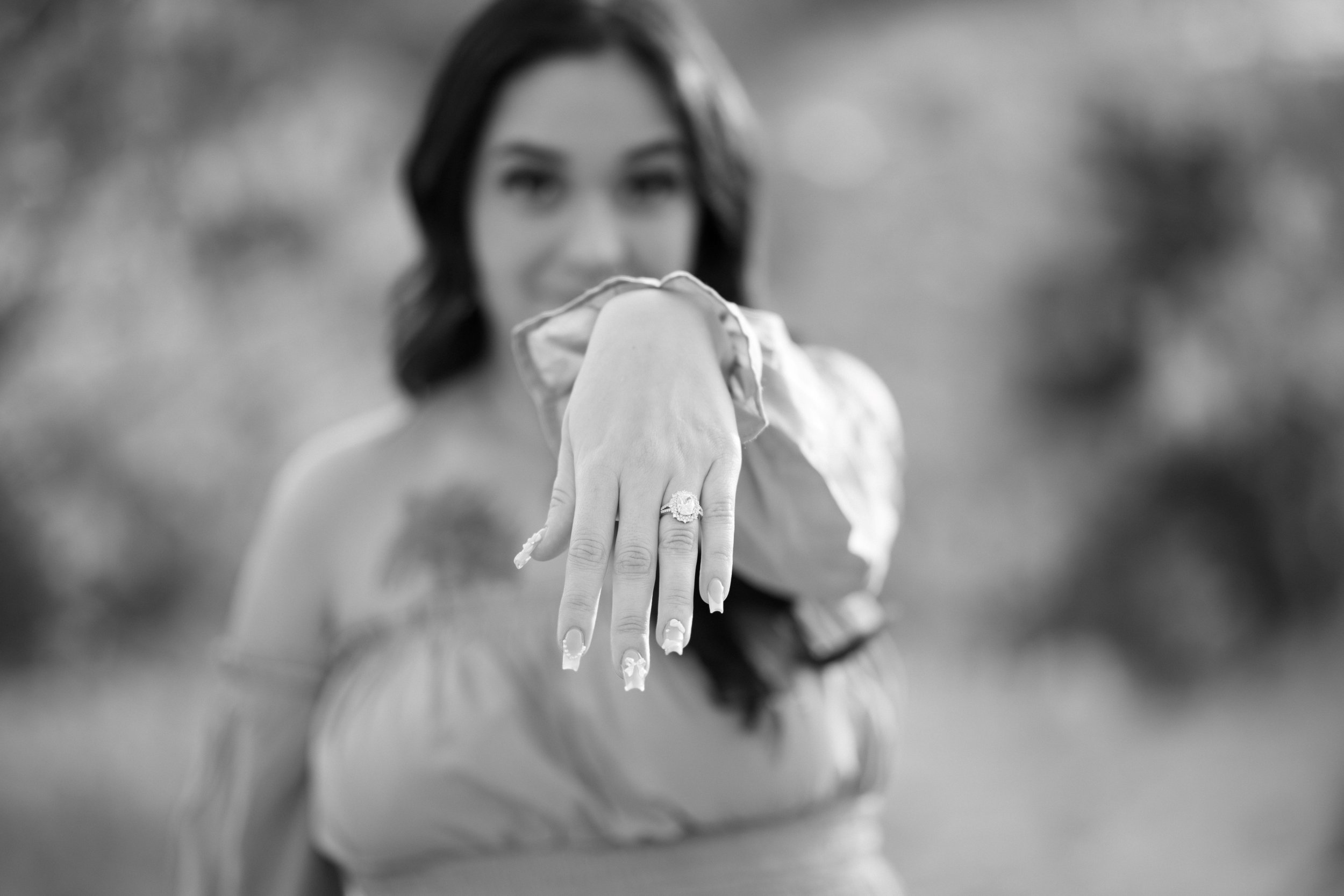 A woman showing her left hand with an engagement ring, with her face out of focus in the background.