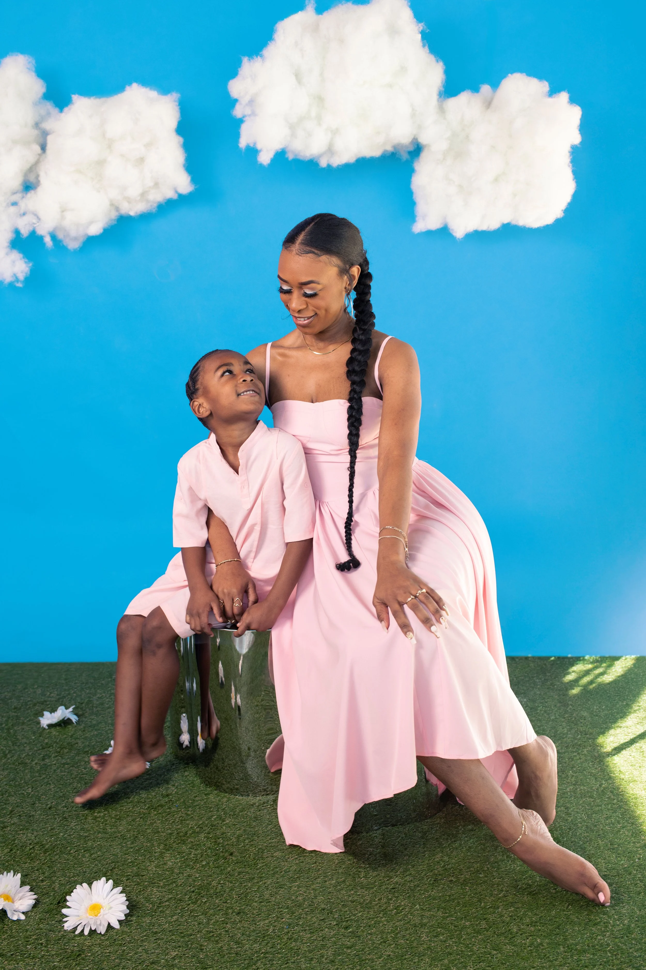 A woman and a young girl sitting together on a metallic stool against a blue background with clouds, both wearing pink dresses. The woman has long braided hair and is looking affectionately at the girl, who is smiling back at her.