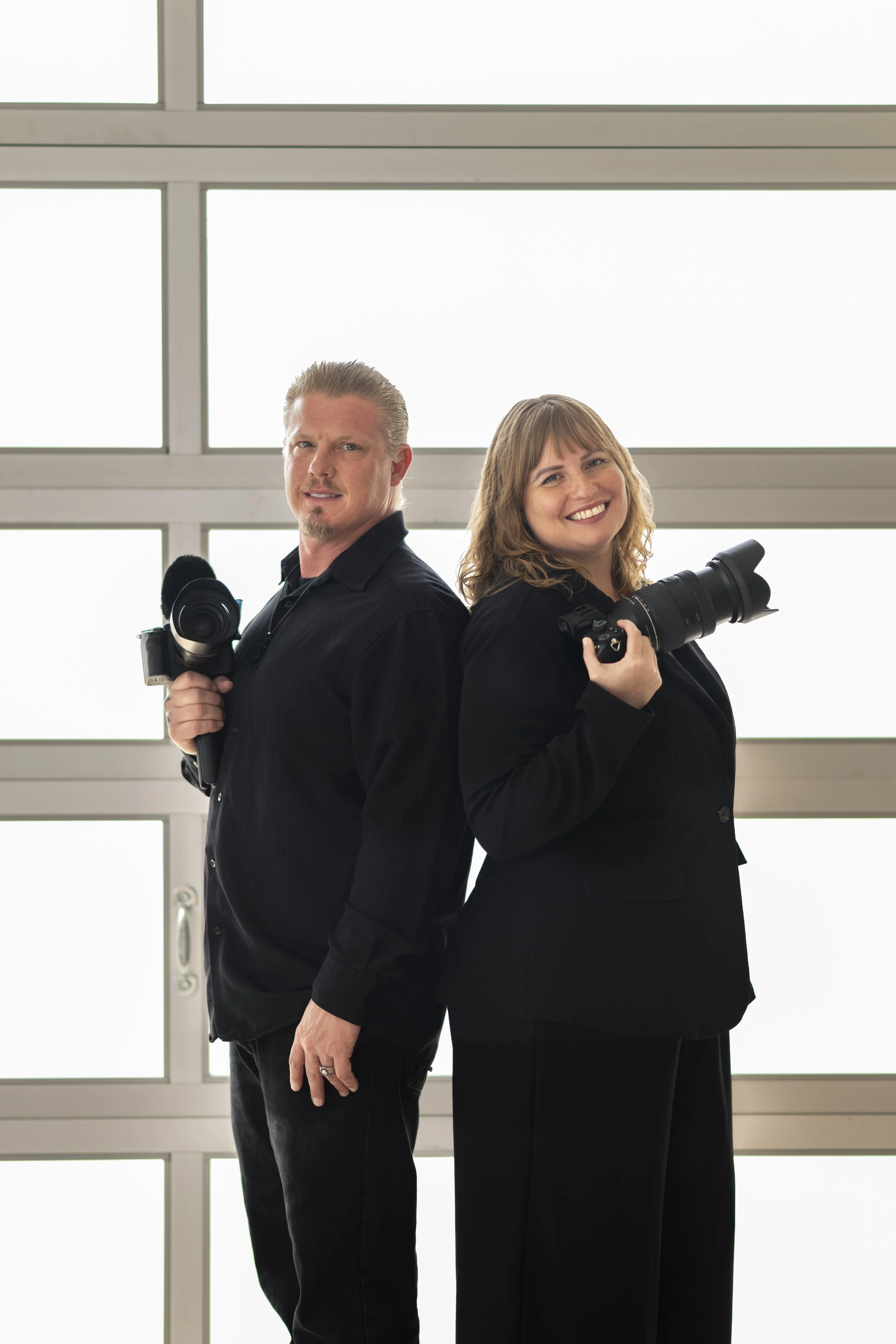 Two people standing back-to-back against a window in a bright room, each holding a camera. The person on the left is a man with short, light-colored hair and a goatee, wearing all black. The person on the right is a woman with shoulder-length, wavy b