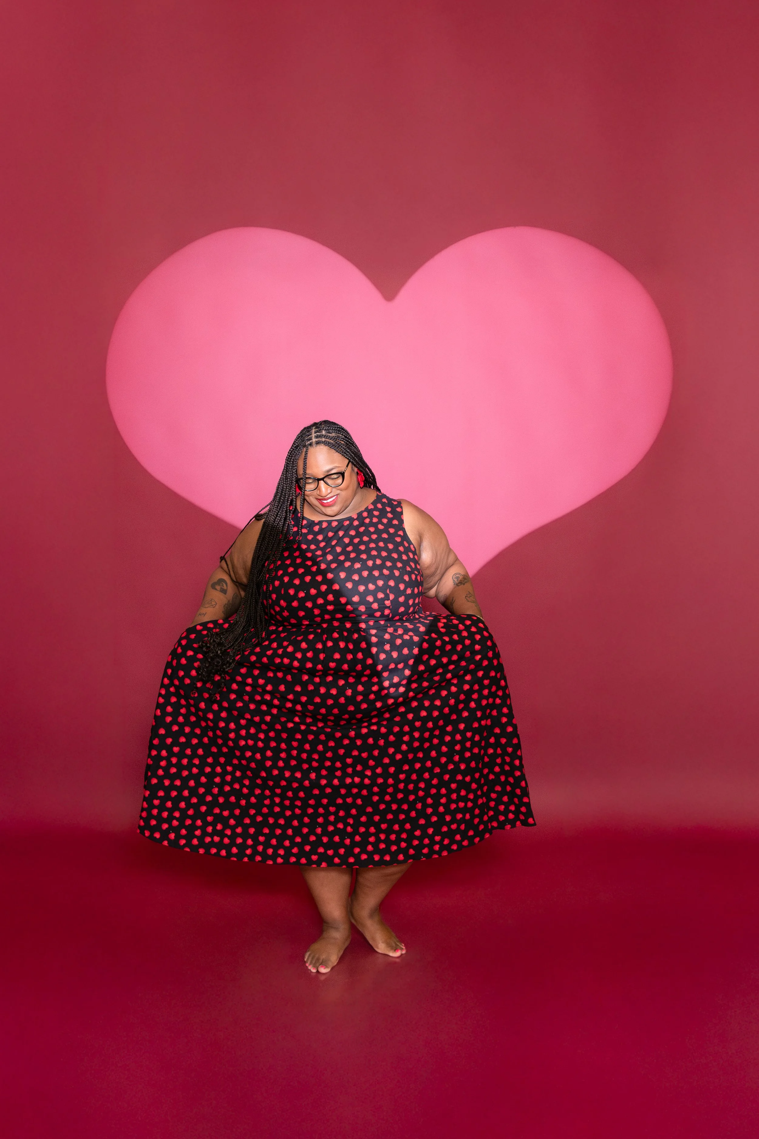 A woman with braided hair and glasses, wearing a black dress with red polka dots, standing barefoot in front of a pink backdrop with a large pink heart.