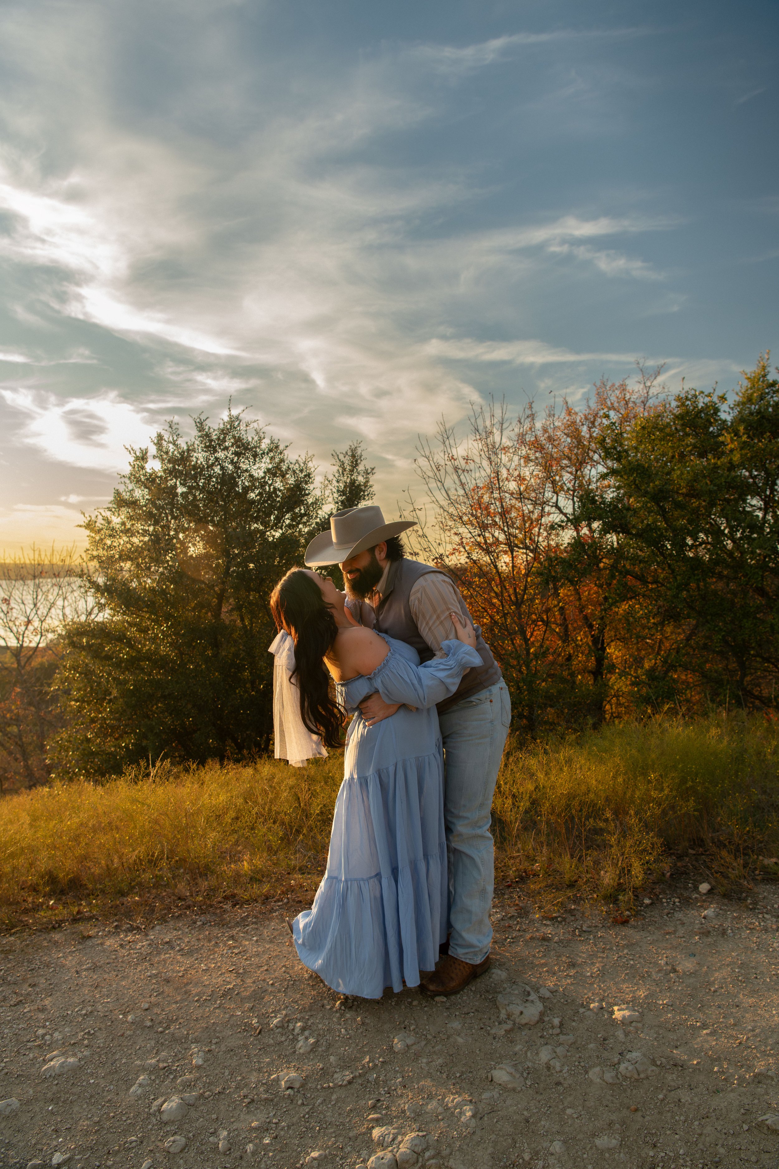 A couple embraces and looks into each other's eyes in a natural outdoor setting during sunset, with trees and a partly cloudy sky in the background.