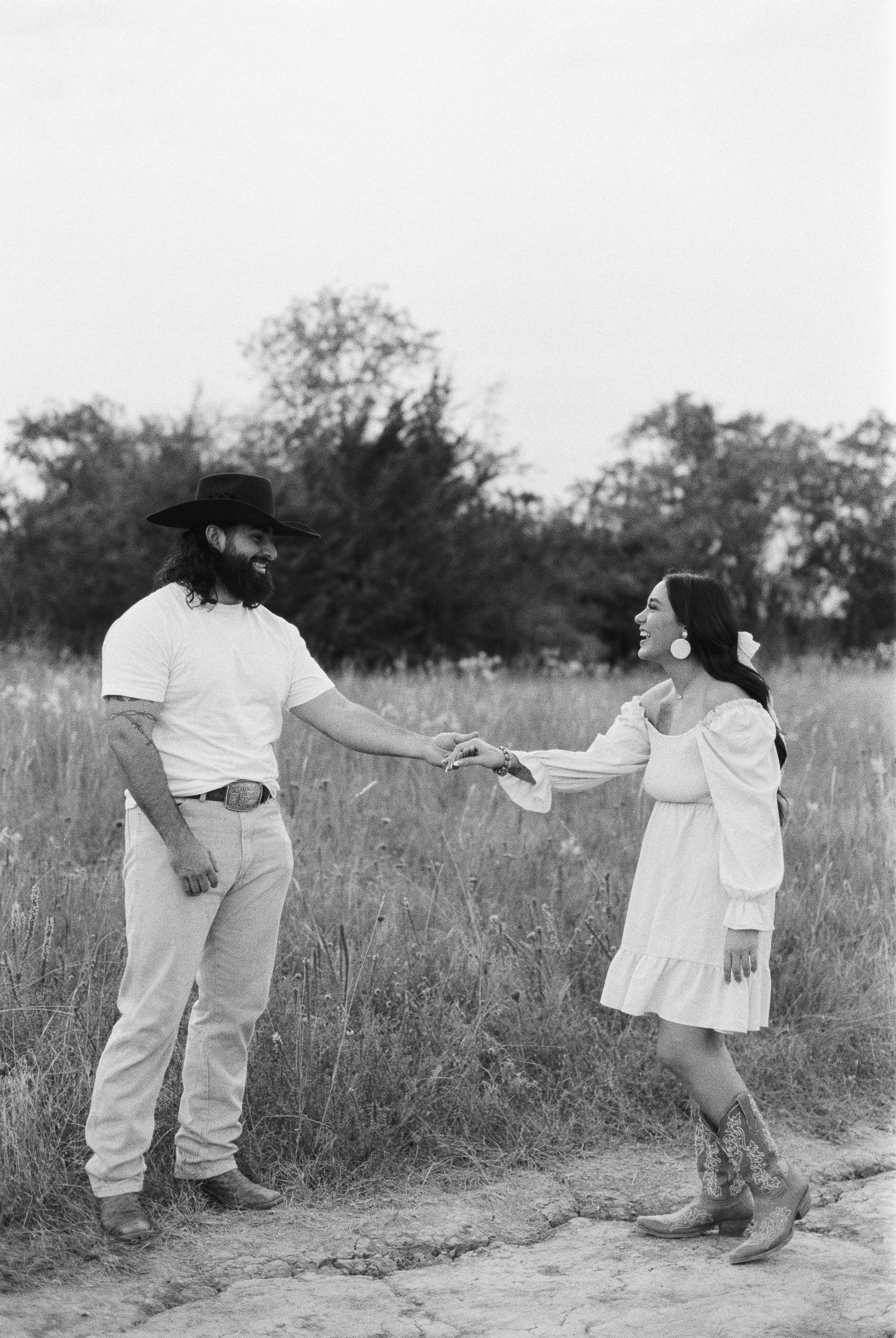 A man and woman holding hands in a field, smiling and laughing, with trees in the background. The man has long hair, a beard, a cowboy hat, a white t-shirt, and light-colored pants. The woman has long dark hair, large earrings, a light-colored dress 