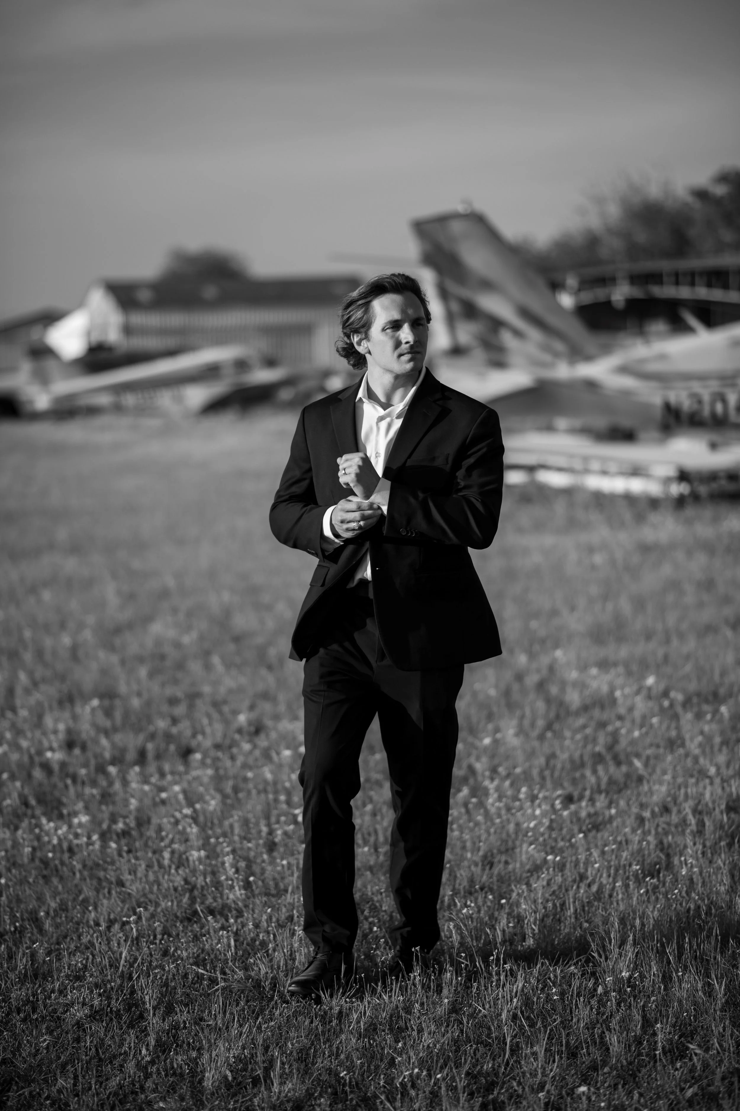 A man in a suit walking on grass with airplanes and hangars in the background, black and white photo.