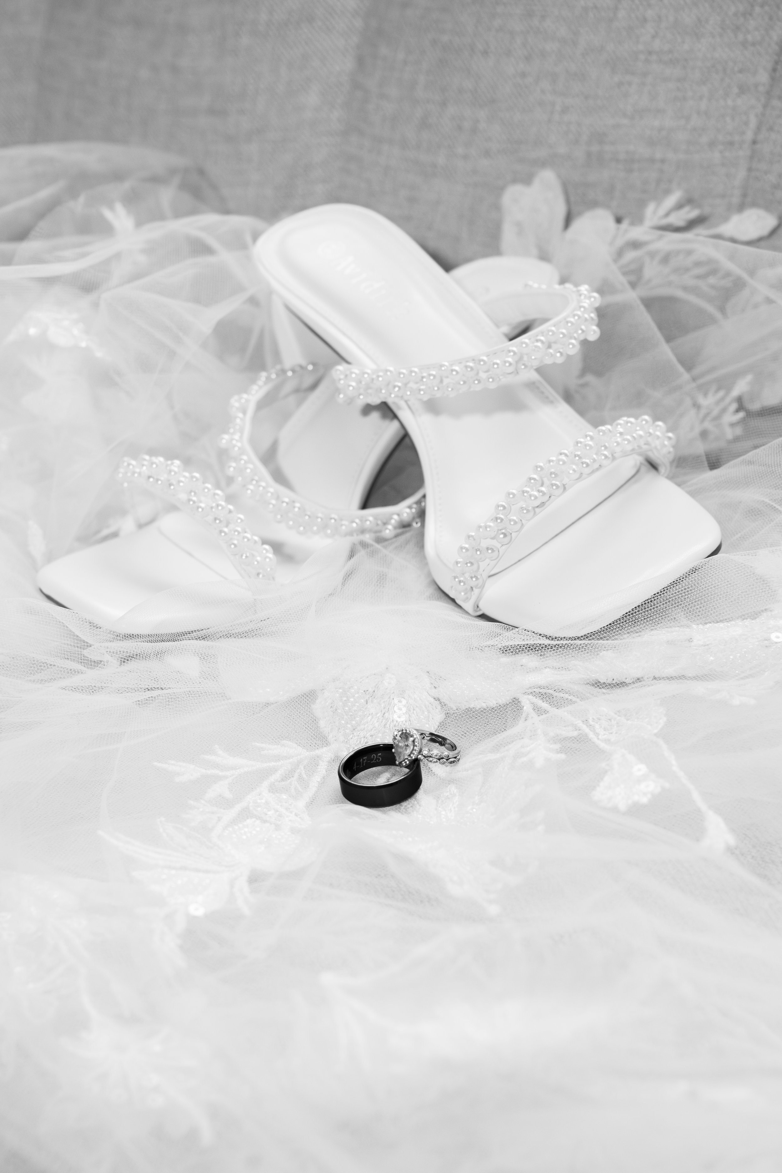 A collection of wedding accessories including a pair of white high-heeled shoes decorated with pearls, a black wedding band, and a wedding ring with a diamond, arranged on lace fabric.