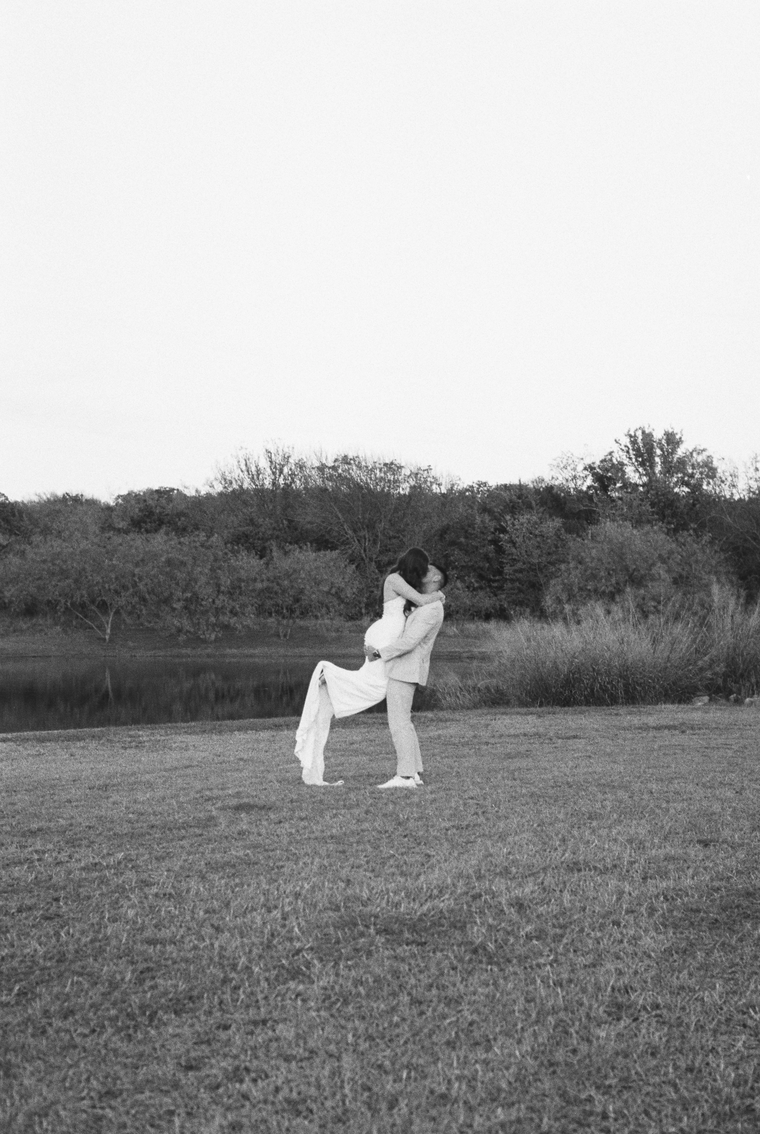 A couple in wedding attire embracing outdoors near a body of water and trees, captured in black and white