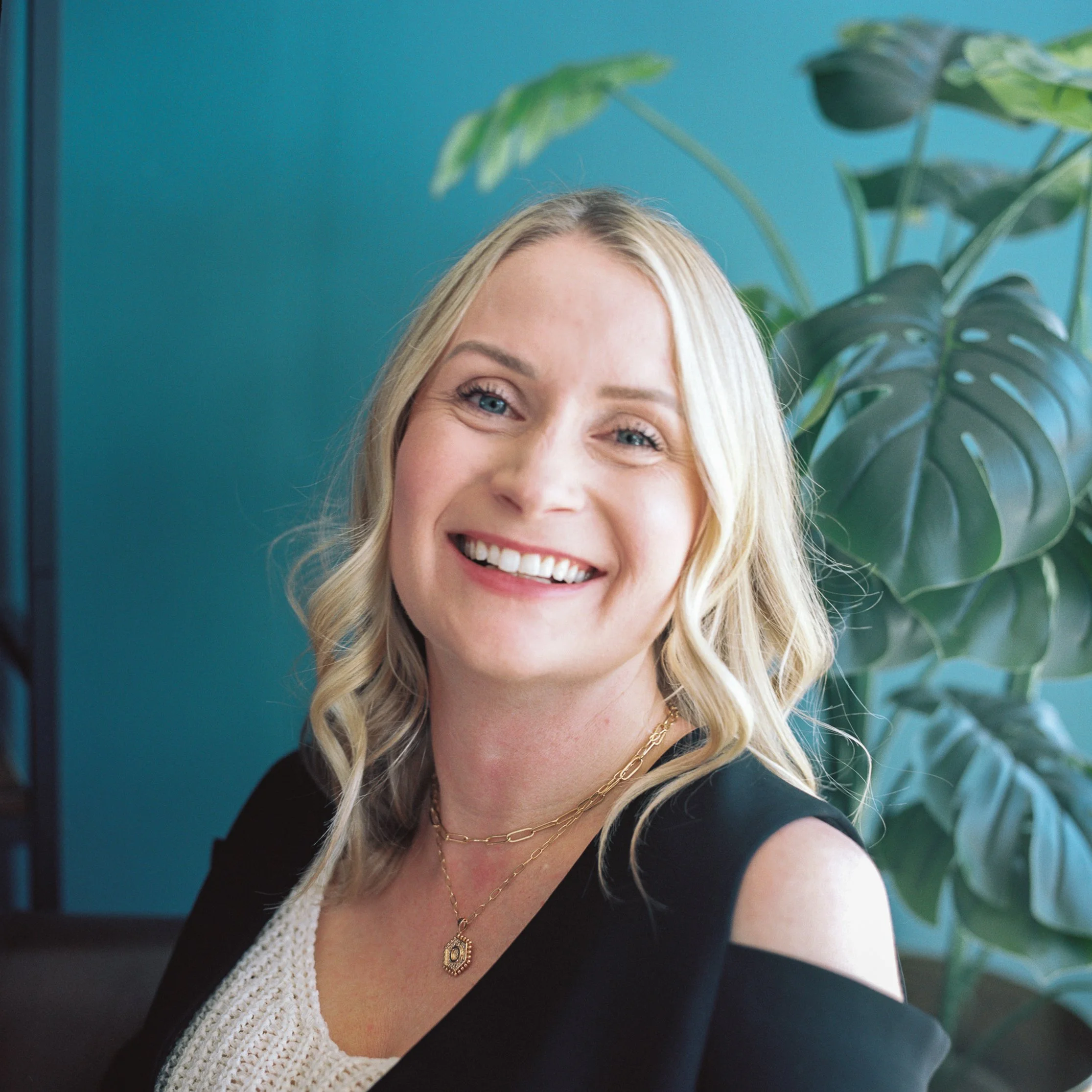 A woman with blonde hair smiling, wearing a black sleeveless top and jewelry, sitting in front of a teal wall and a large green plant.