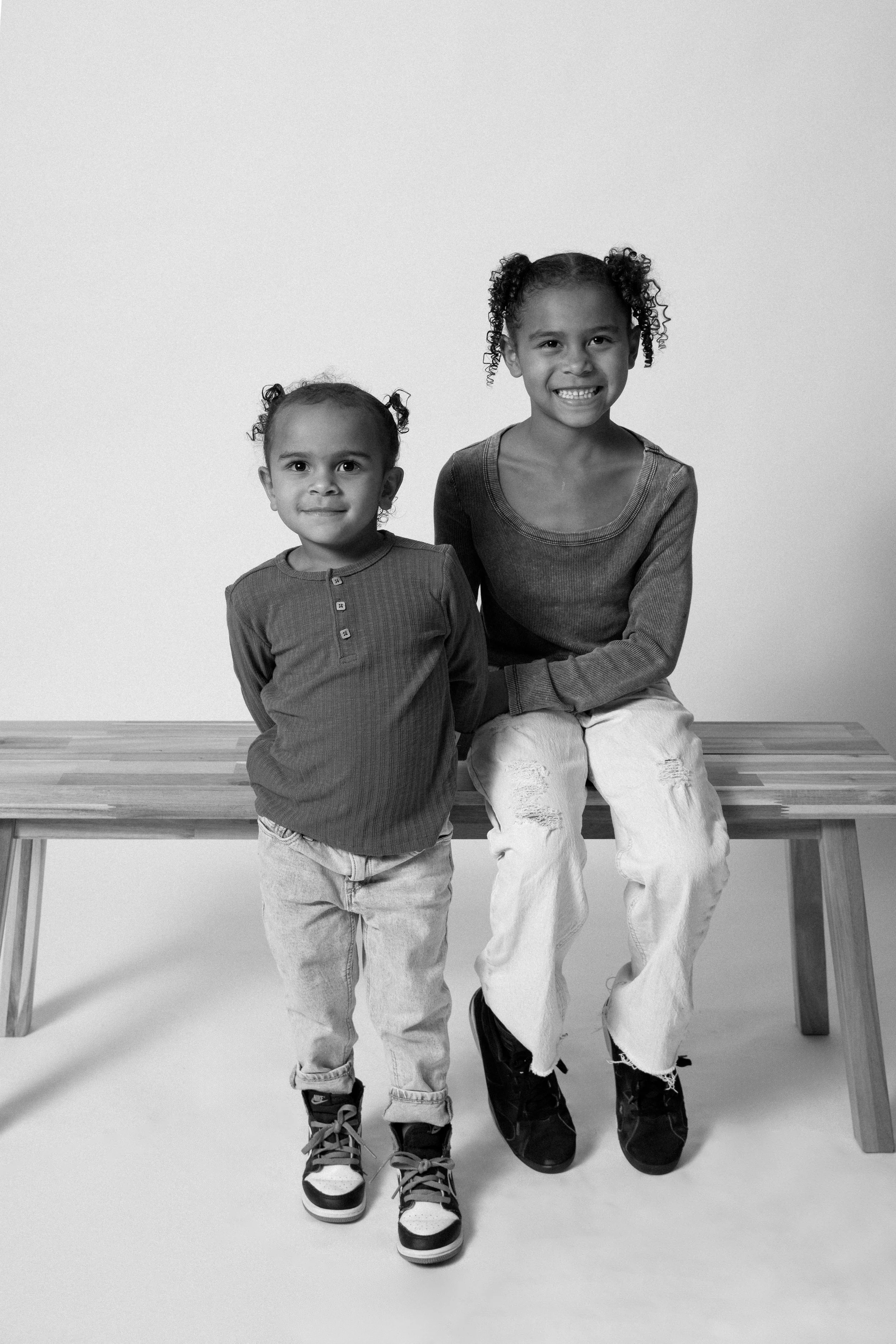 Two young girls, one sitting on a bench and the other standing beside her, both smiling in a black-and-white photo.
