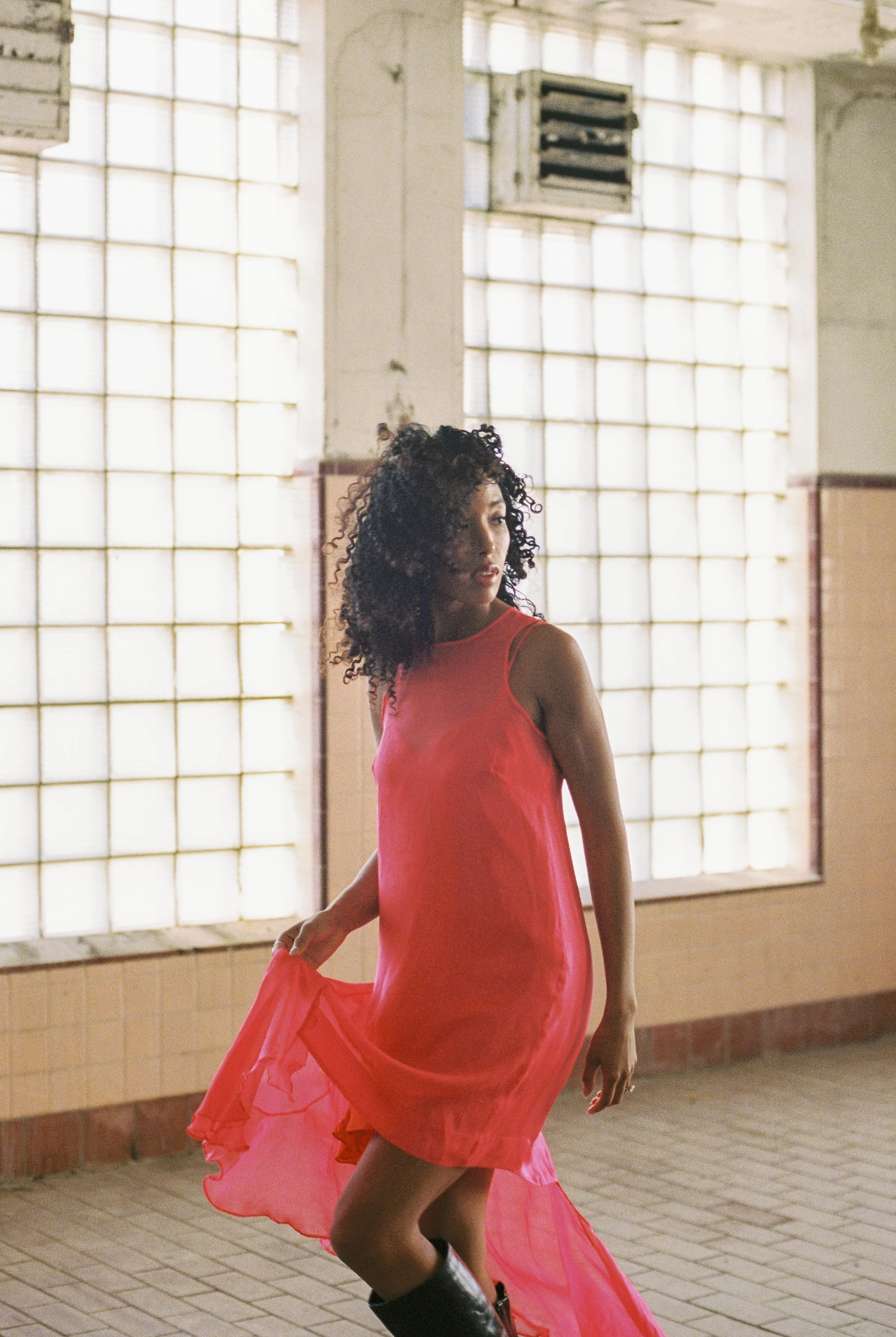 A woman with curly hair, wearing a flowing pink dress and black boots, dancing in an industrial-style room with large glass block windows.