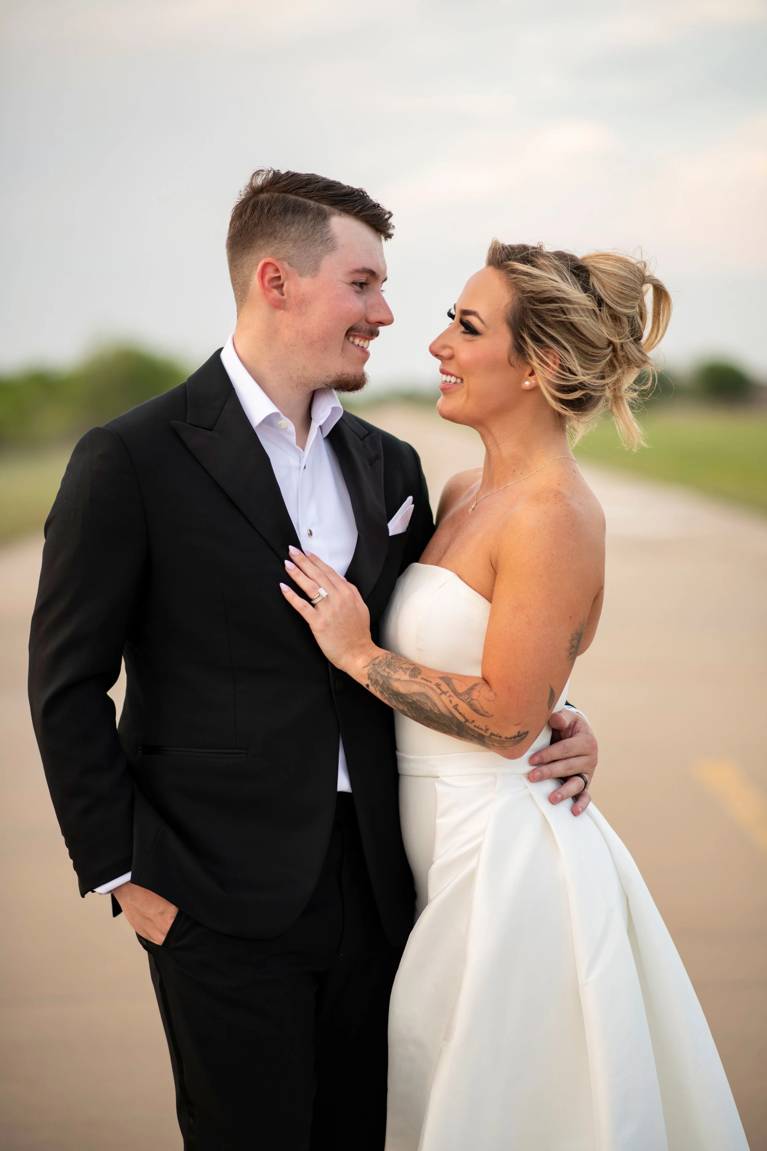 A couple dressed in wedding attire, standing close together outdoors with a blurred background of green trees and an overcast sky, smiling and gazing into each other's eyes.
