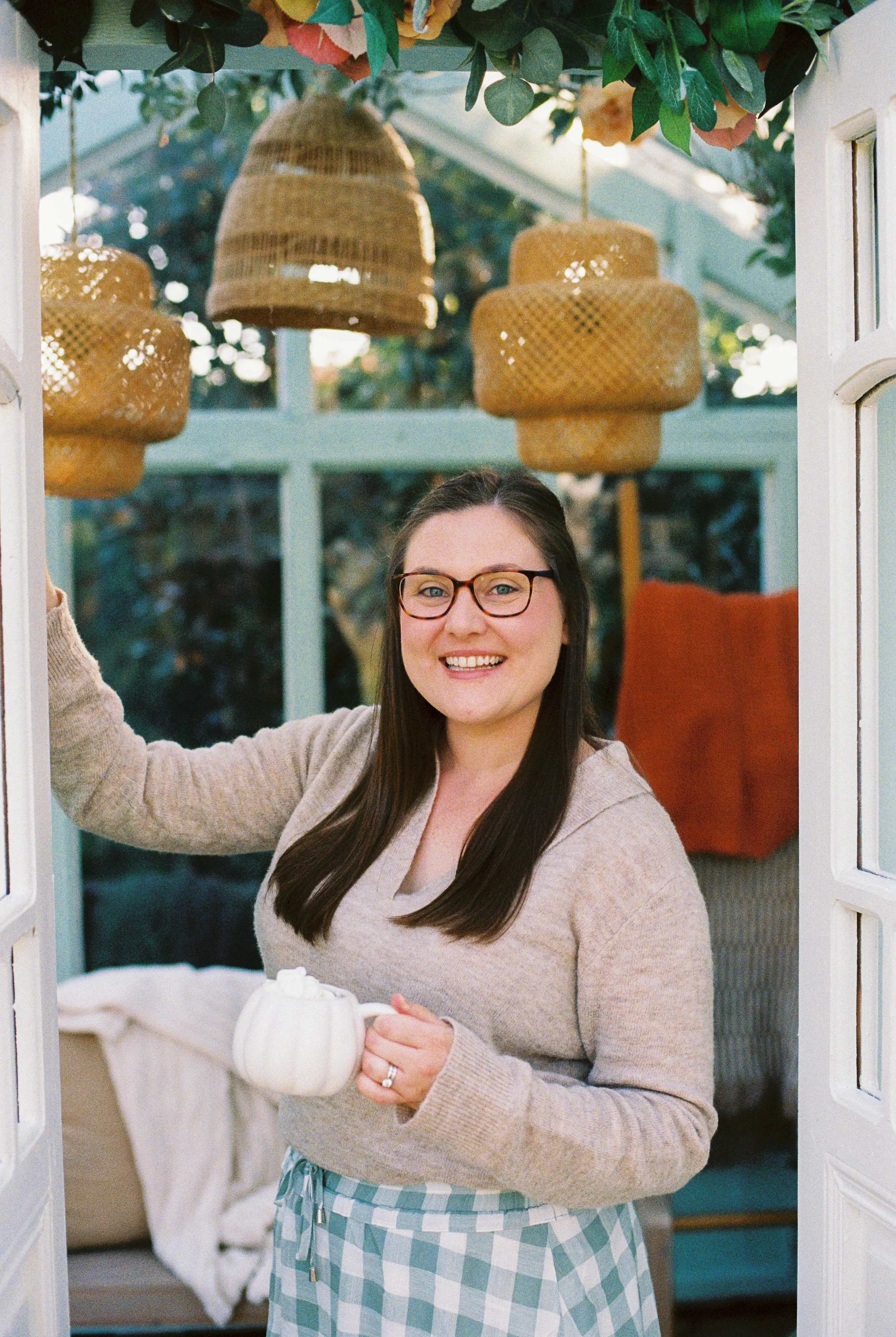 A woman with dark hair, glasses, and light-colored sweater holding a white mug standing in a sunroom, smiling at the camera. The sunroom has hanging wicker light fixtures and greenery on the ceiling, with a cozy seating area in the background.