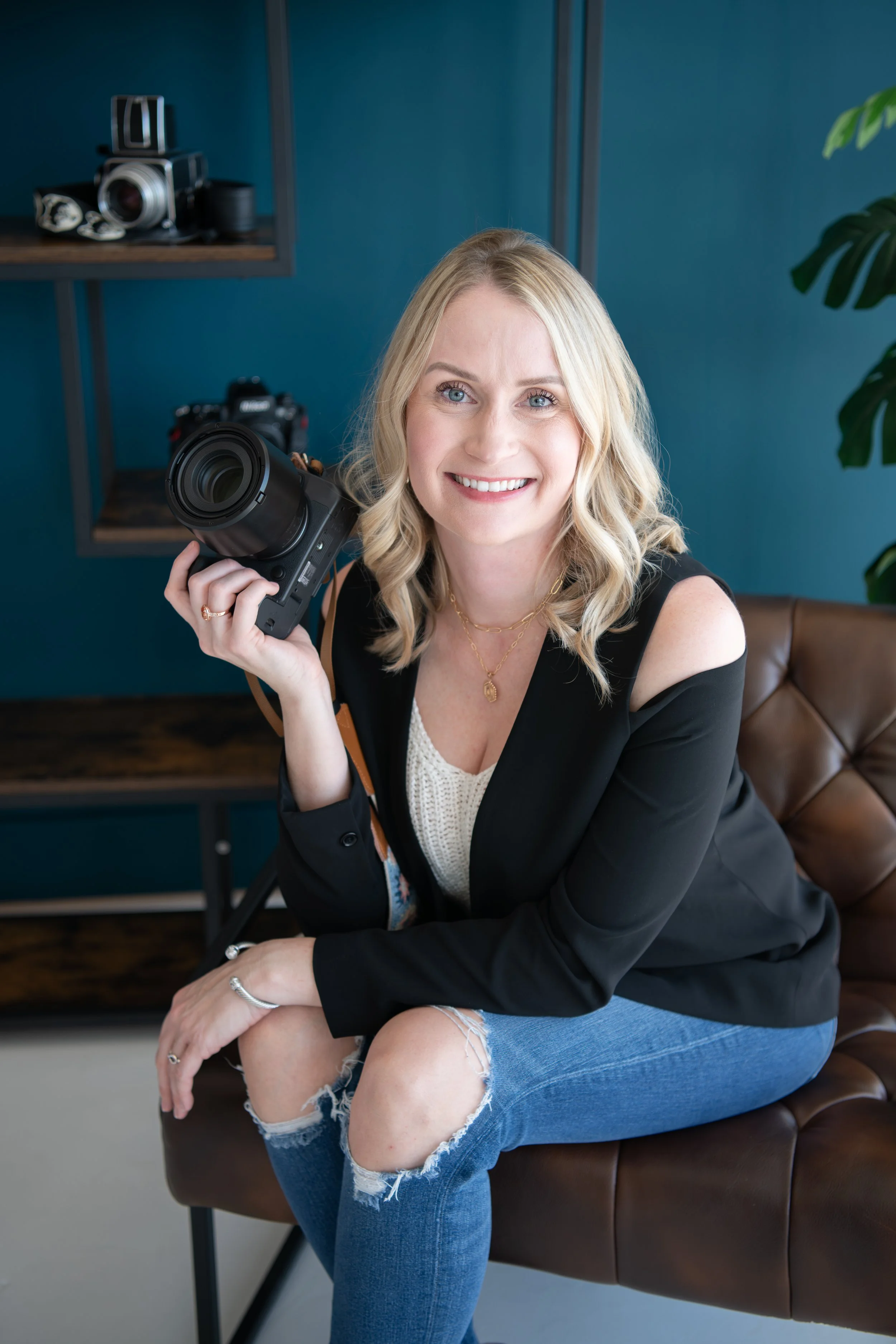 A woman with blonde hair smiling while holding a professional camera, seated on a brown leather chair in a modern room with blue walls and shelves with cameras and photography equipment.
