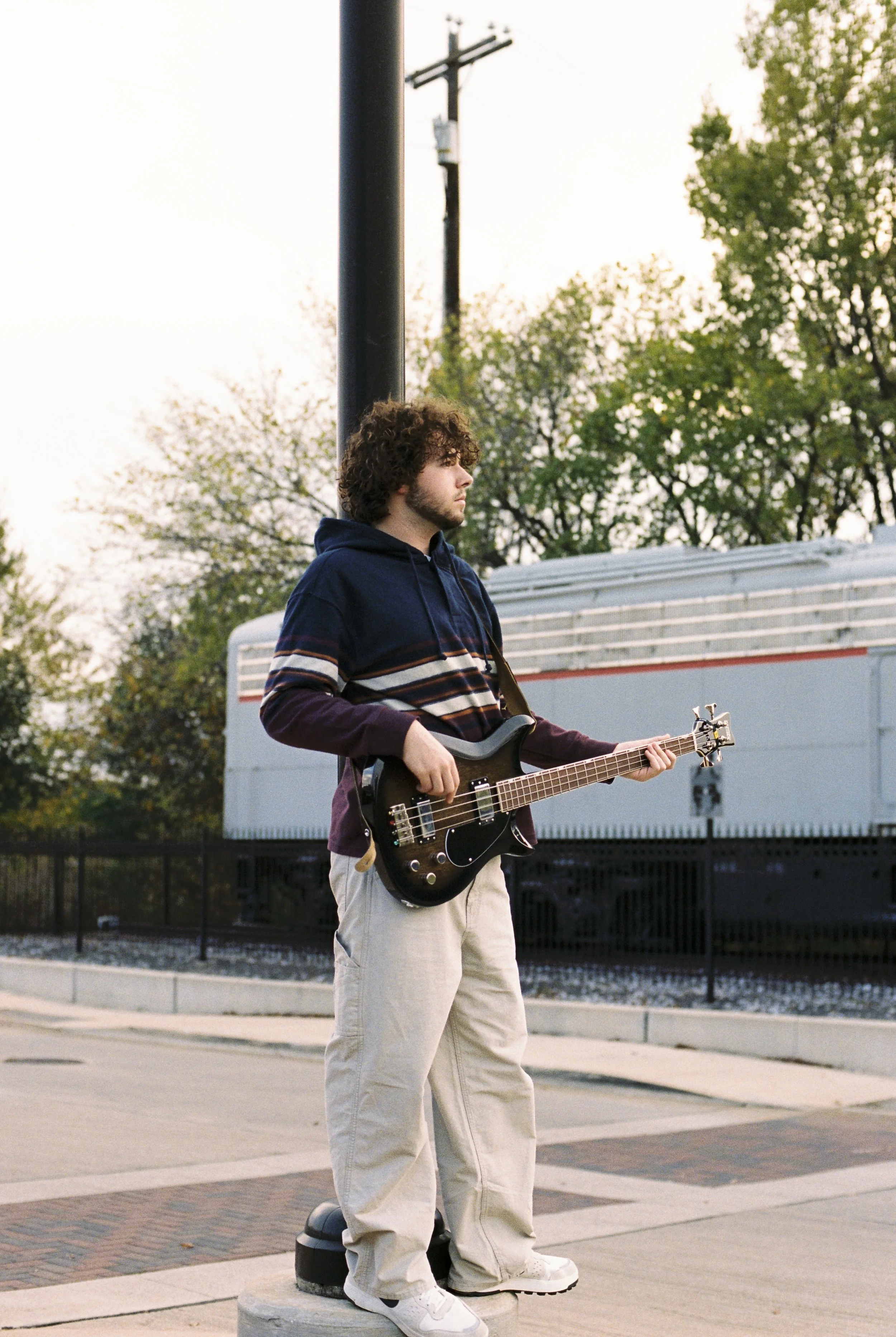 A young man with curly hair and a beard standing on a concrete block, playing a black electric guitar outdoors during sunset, with trees, utility pole, and a train in the background.