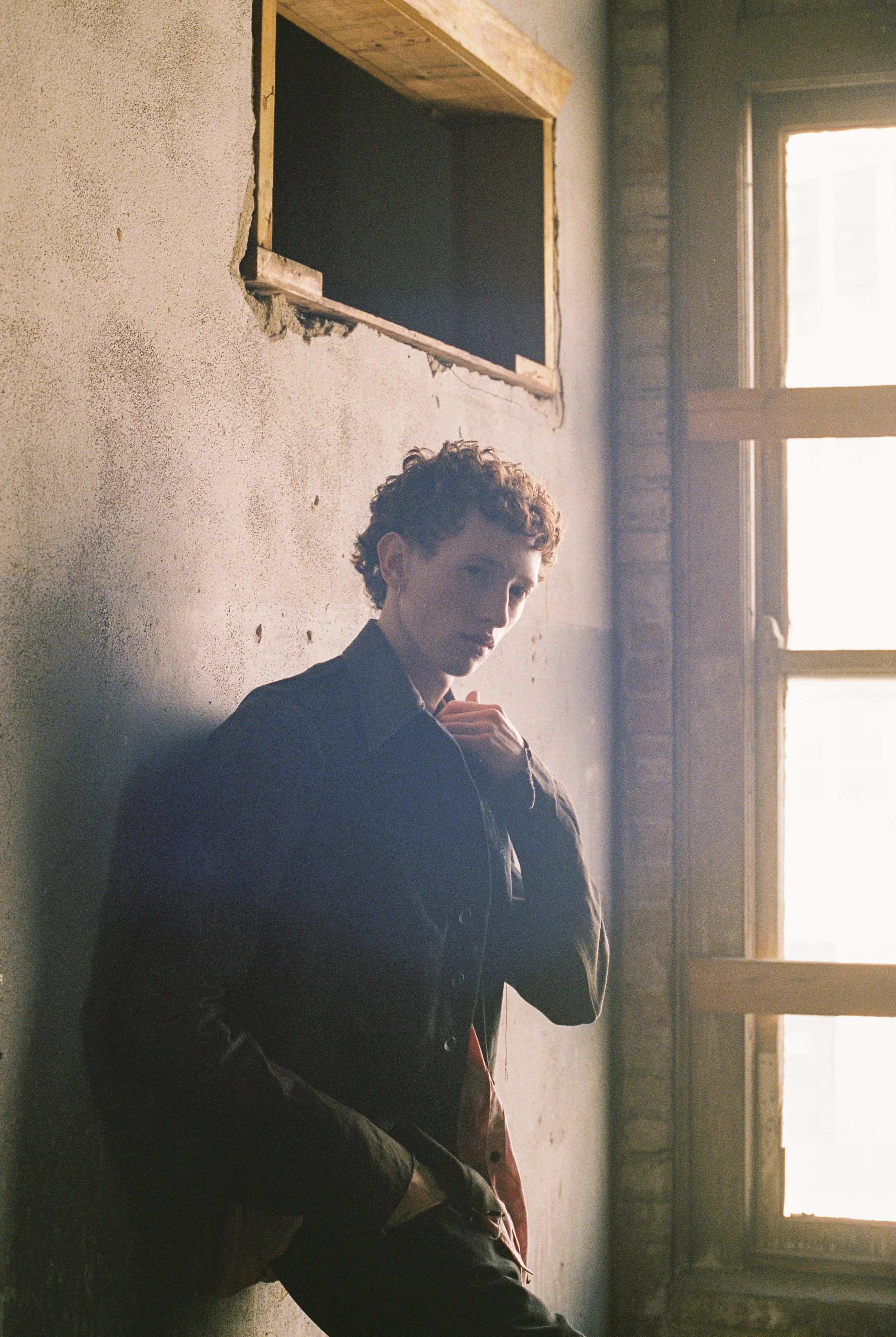 A person with short curly hair and a dark jacket sitting on the floor near a wall with a window in the background, which lets in natural light.