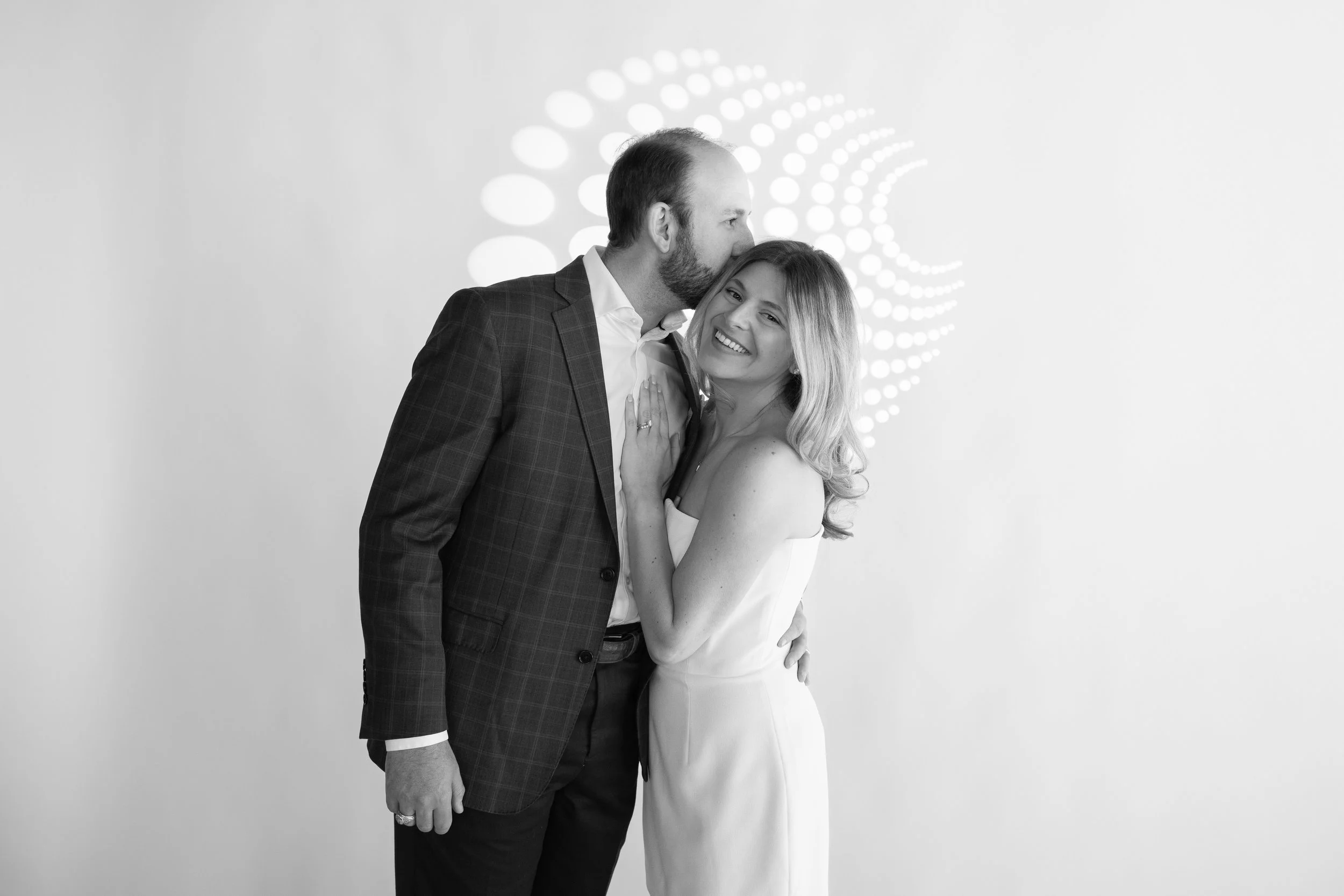 A black and white photo of a happy couple. The man is kissing the woman on the forehead, and she is smiling. The woman is wearing a strapless dress, and the man is dressed in a suit jacket. The background features a glowing, circular pattern.