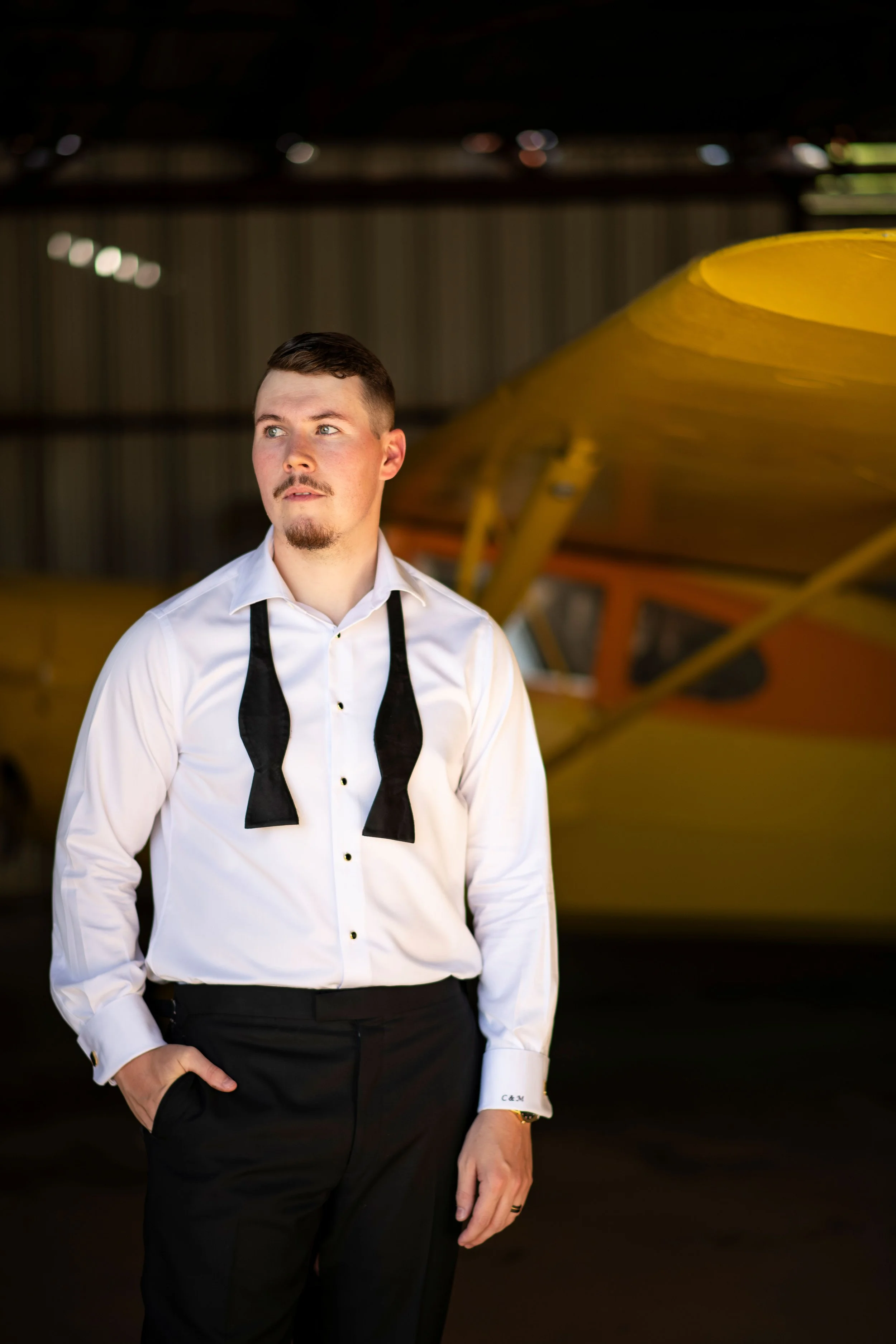 A man dressed in formal black and white attire standing inside an airplane hangar, with a yellow airplane in the background.