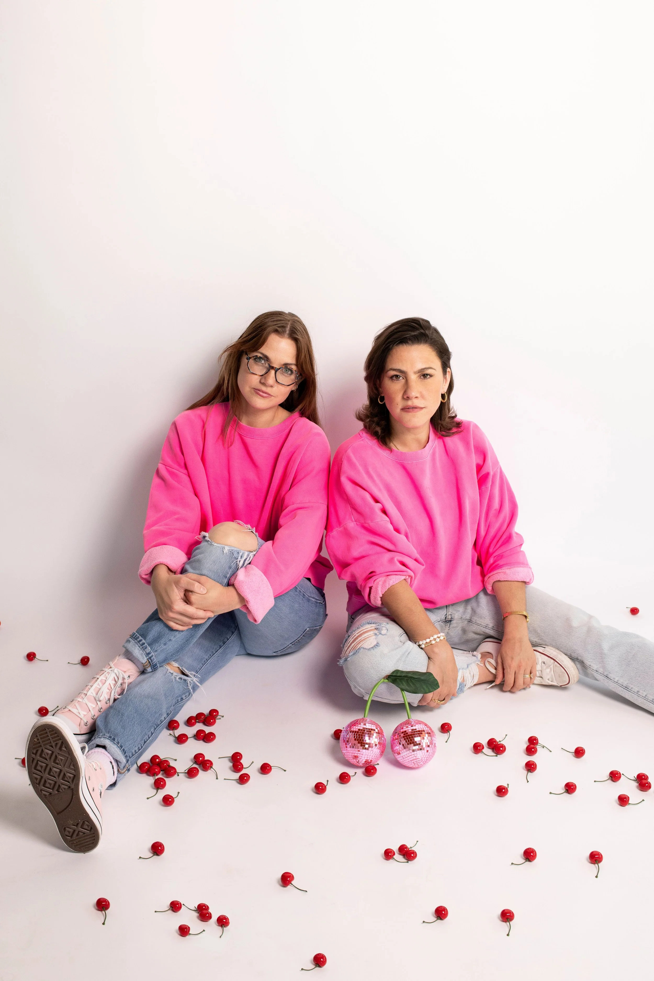 Two women wearing pink sweaters sitting on the floor surrounded by scattered cherries, with two pink disco balls shaped like cherries in front of them.