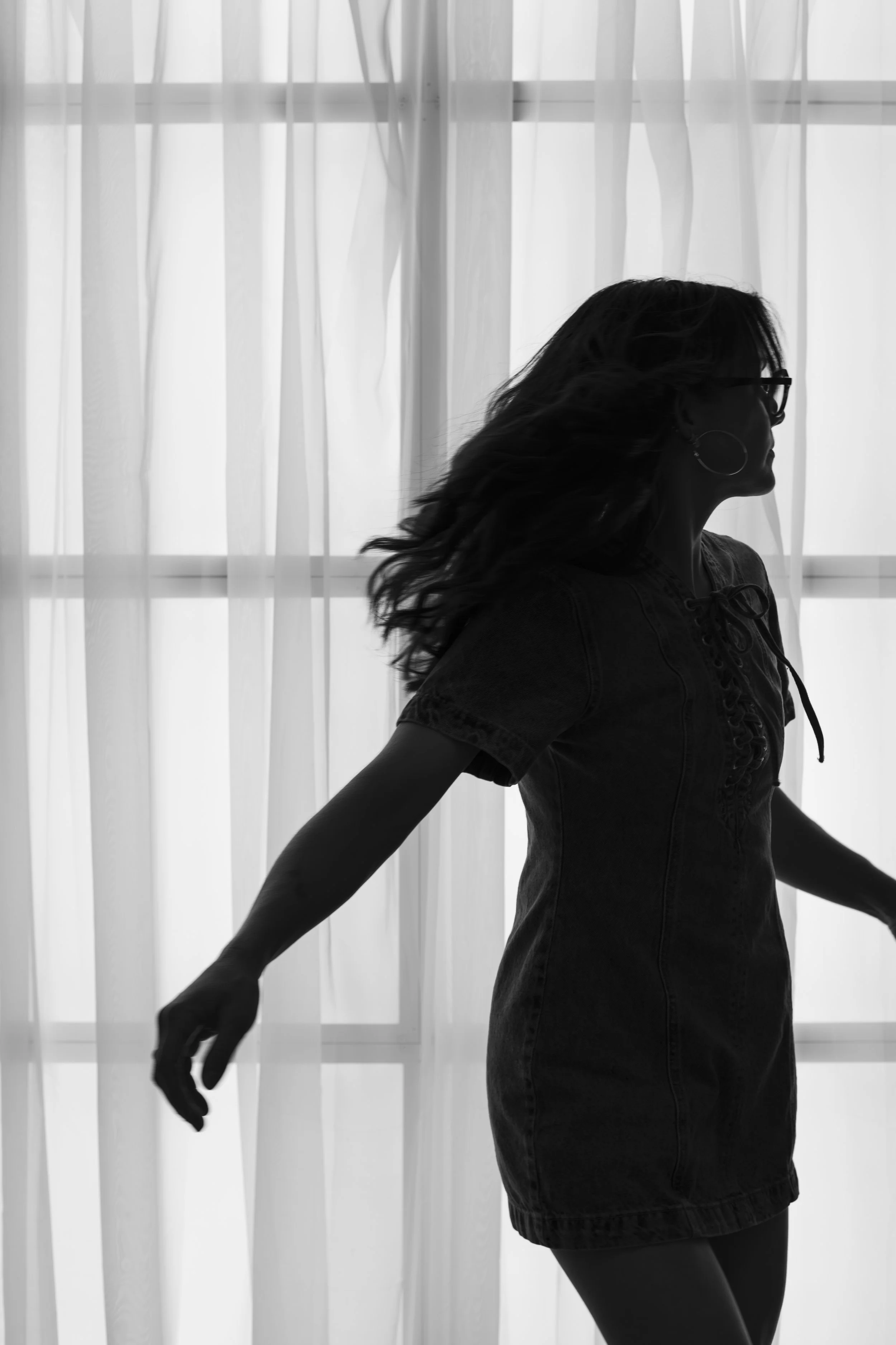 Silhouette of a woman with wavy hair wearing glasses and hoop earrings, standing in front of sheer curtains.