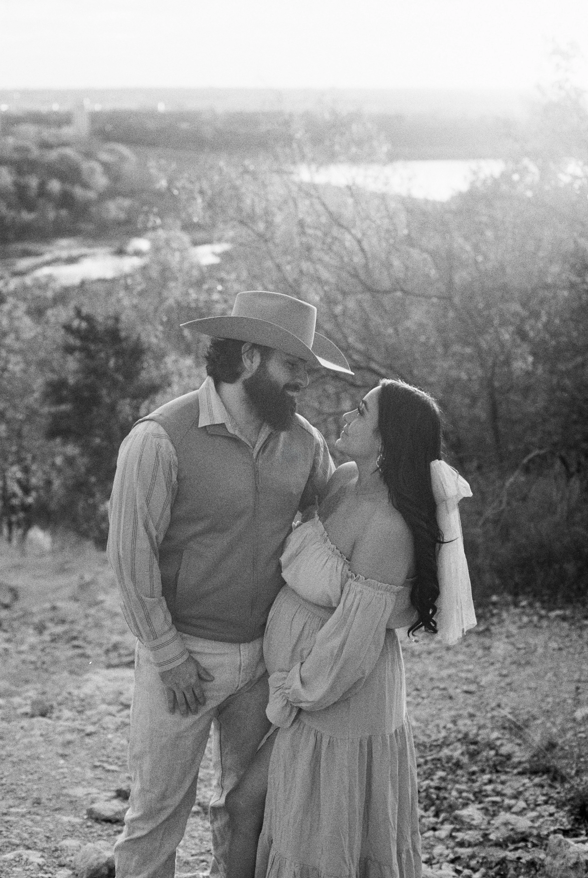 A black-and-white photo of a man with a beard wearing a cowboy hat and a vest, and a woman with long dark hair in a flowing off-shoulder dress, standing close together outdoors with a scenic landscape in the background.