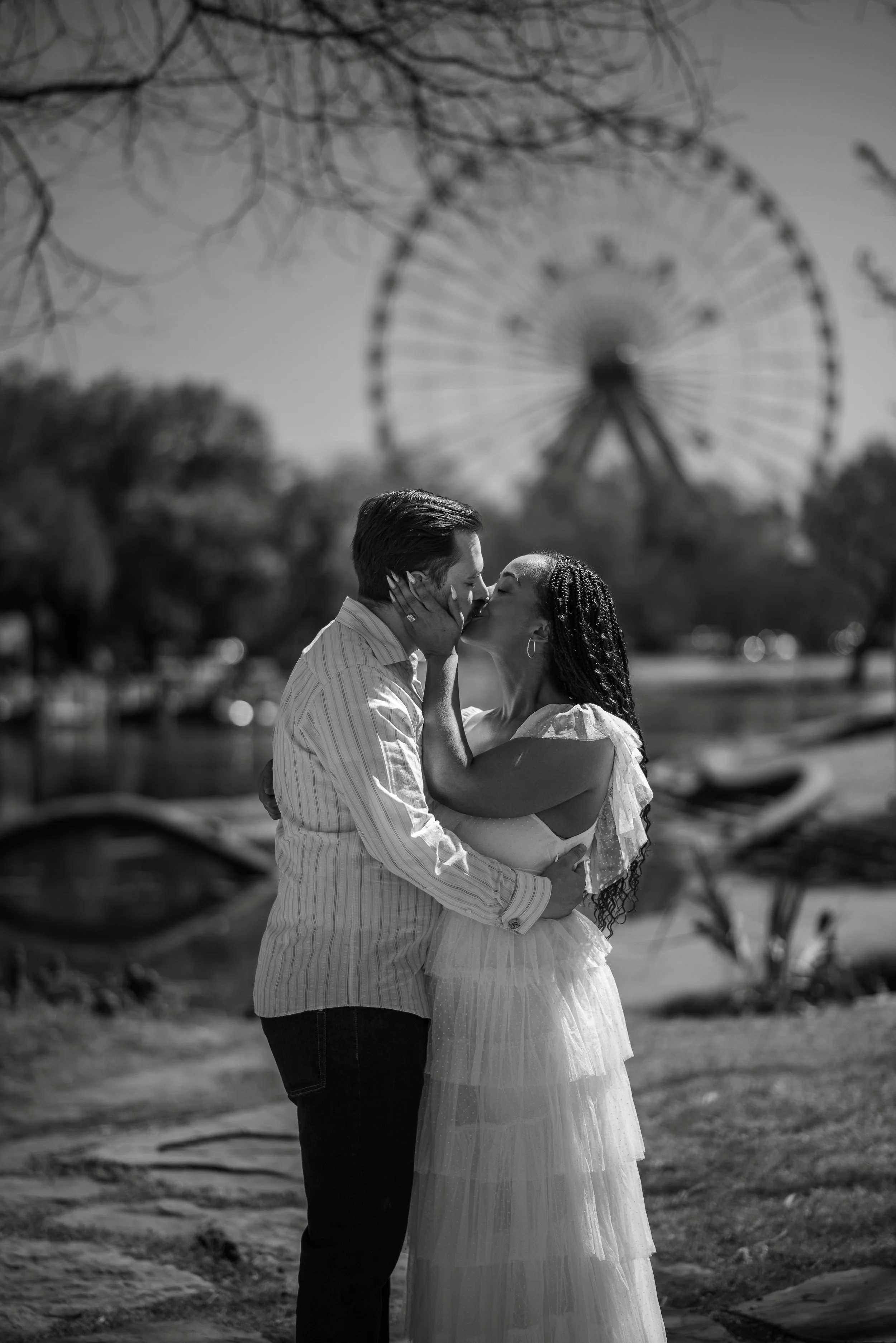 A couple kissing outdoors near a lake with a Ferris wheel in the background, in black and white.