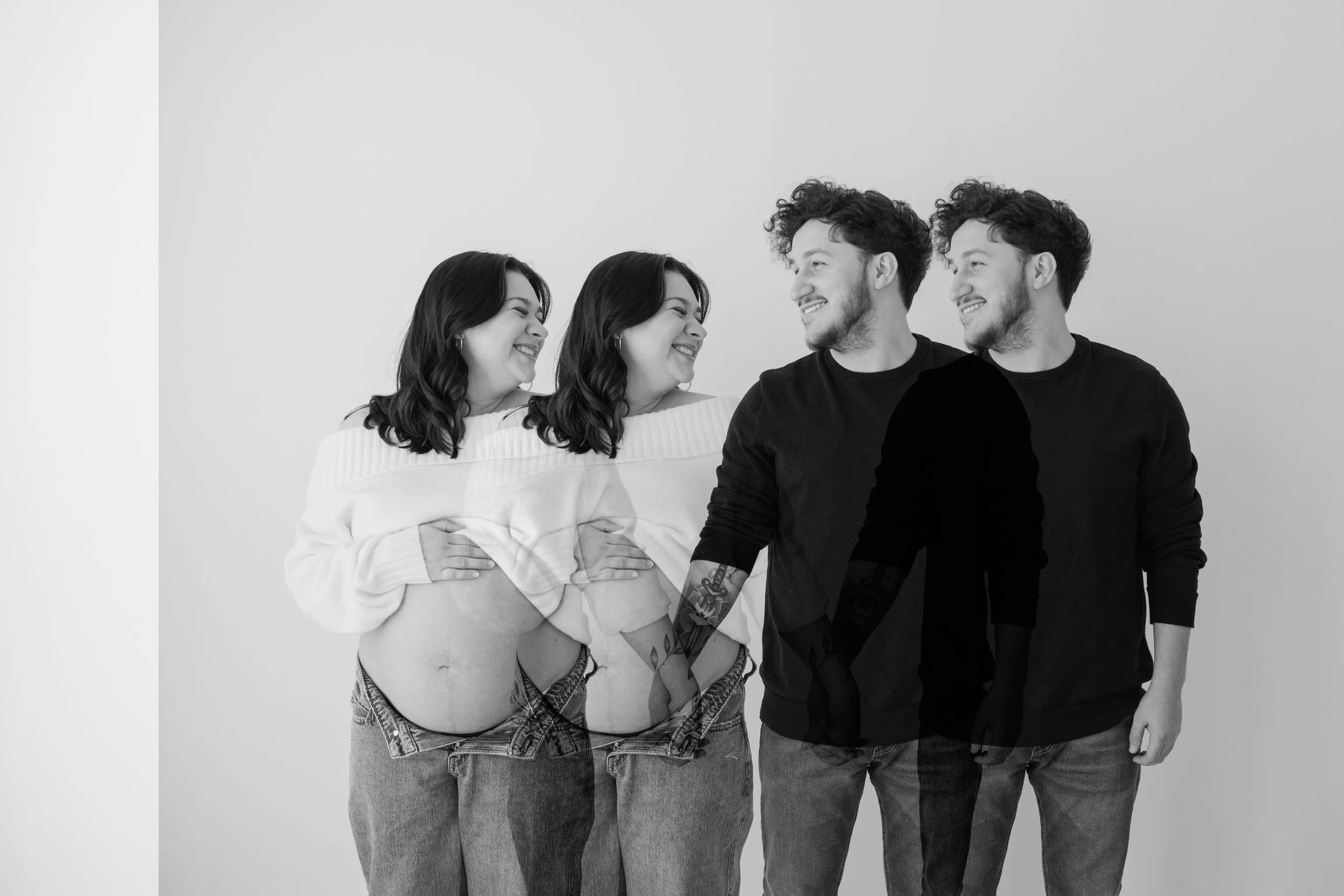 Black and white photo of a woman and a man looking at each other, smiling, with the woman lifting her shirt to show her pregnant belly, and the man and woman holding hands.
