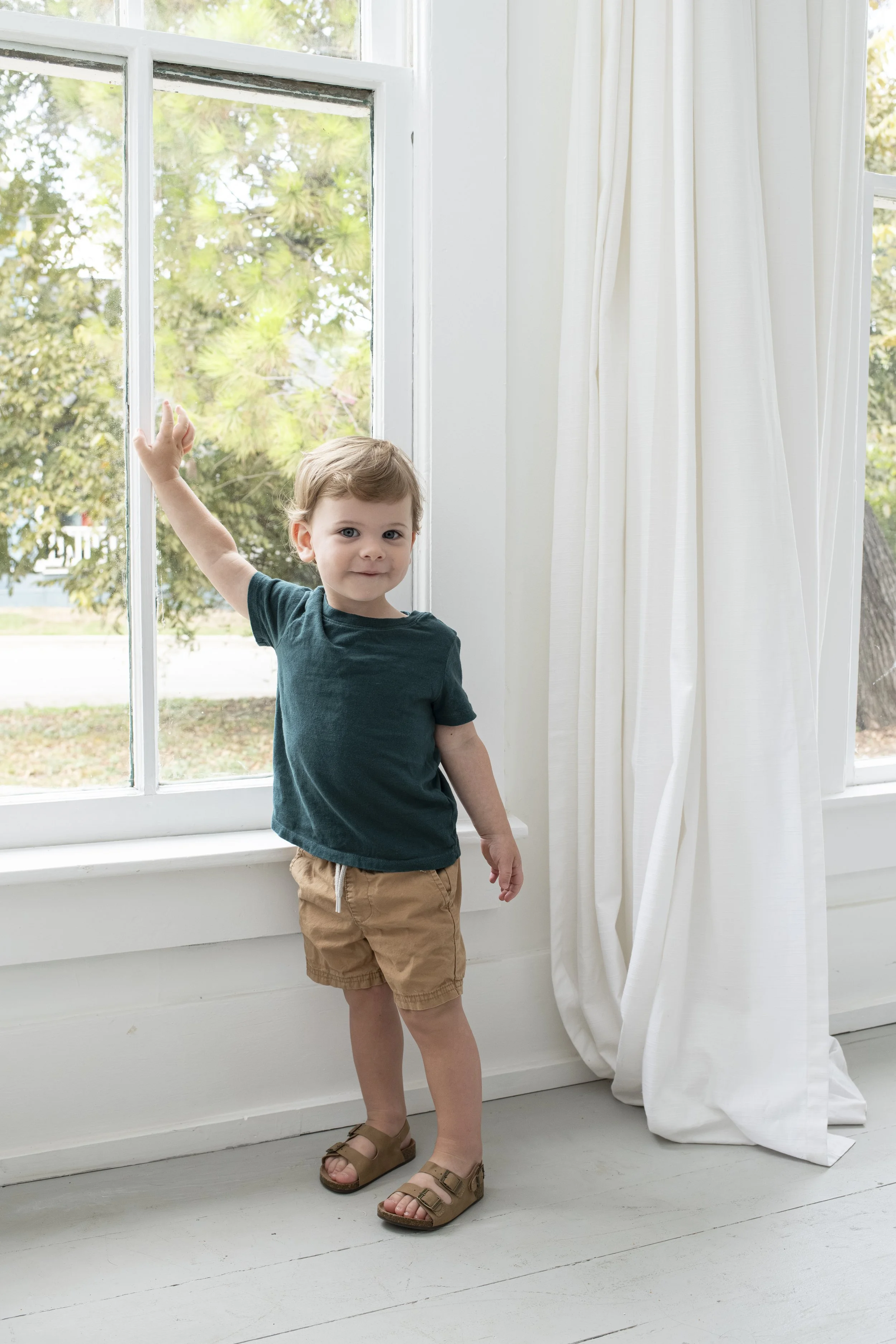 A young boy stands inside near a large window with white frames and white curtains, looking at the camera while resting his arm on the window frame. Outside, trees and part of a sidewalk are visible.