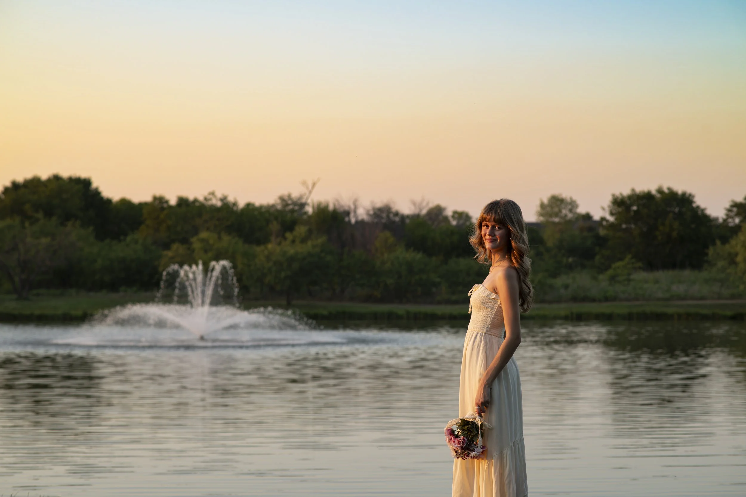 A woman in a beige dress holding a bouquet of flowers standing near a lake with a fountain, trees in the background, during sunset.