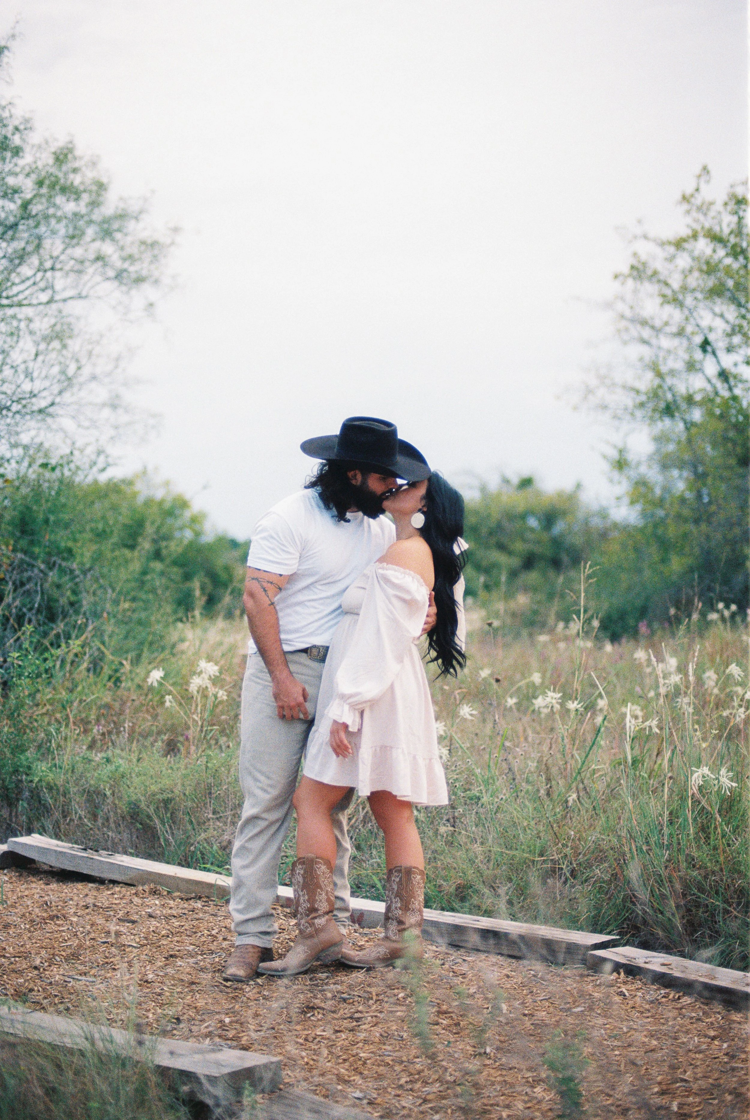 A man and woman kiss in a field. The man wears a black cowboy hat, white t-shirt, and light gray pants. The woman wears a light pink dress and cowboy boots. They stand on a dirt path with greenery and white flowers around them.