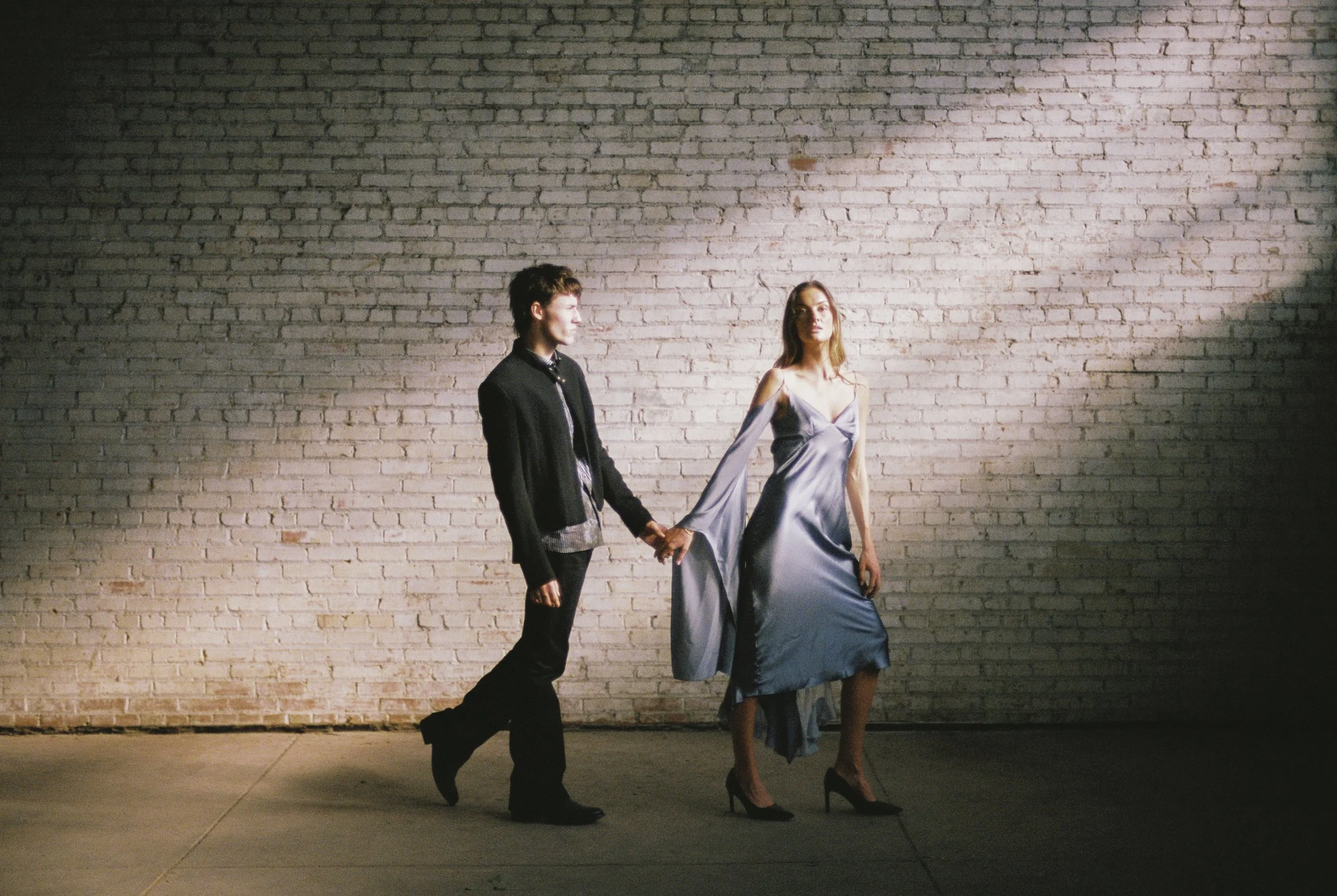 A man and a woman holding hands in front of a brick wall. The woman is wearing a silky, off-shoulder dress and high heels. The man is dressed in a suit jacket and dark pants. The scene appears to be outdoors, possibly at night, with dramatic lighting