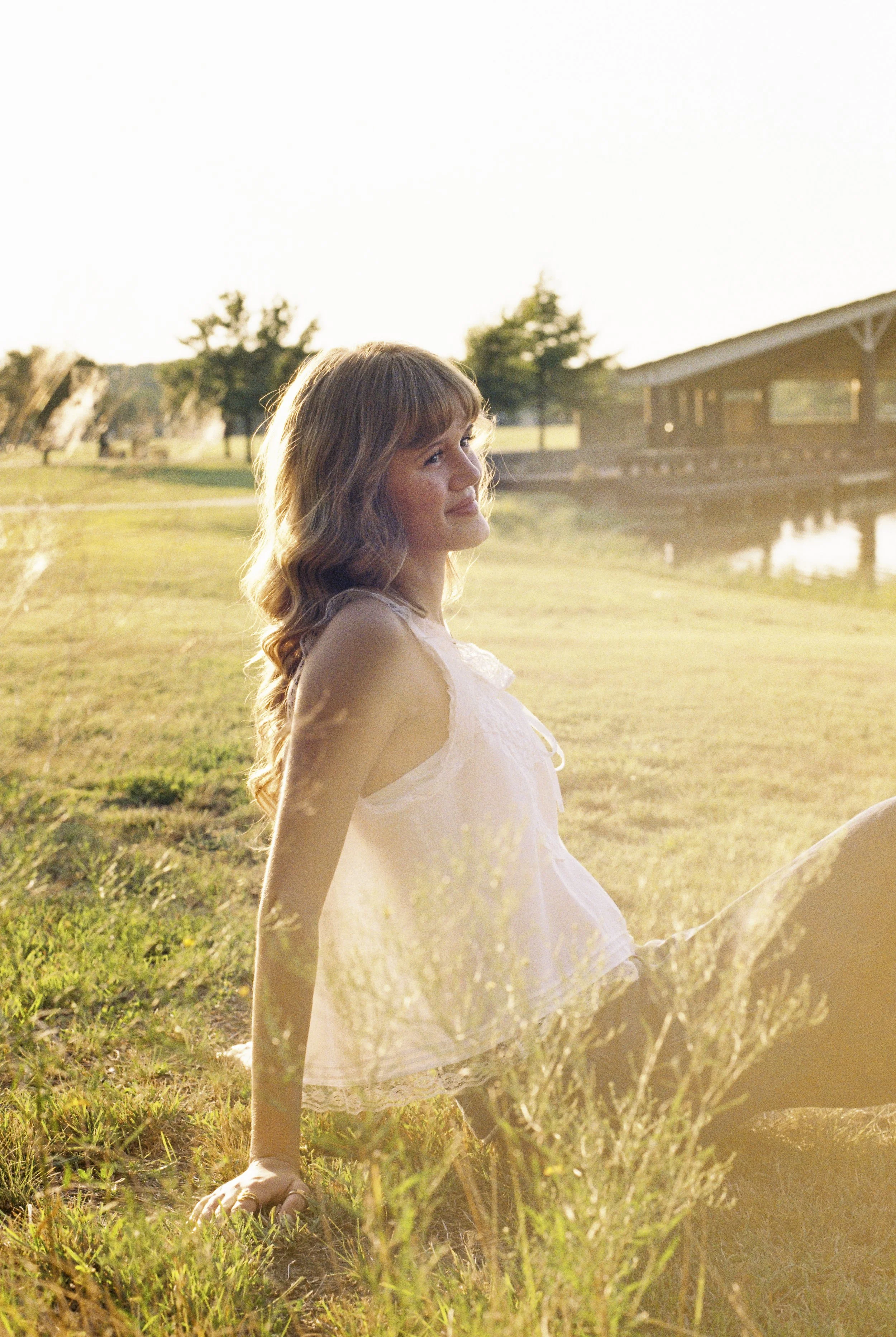 A young woman sitting on grass near a pond, smiling, with trees and a building in the background, during sunset.