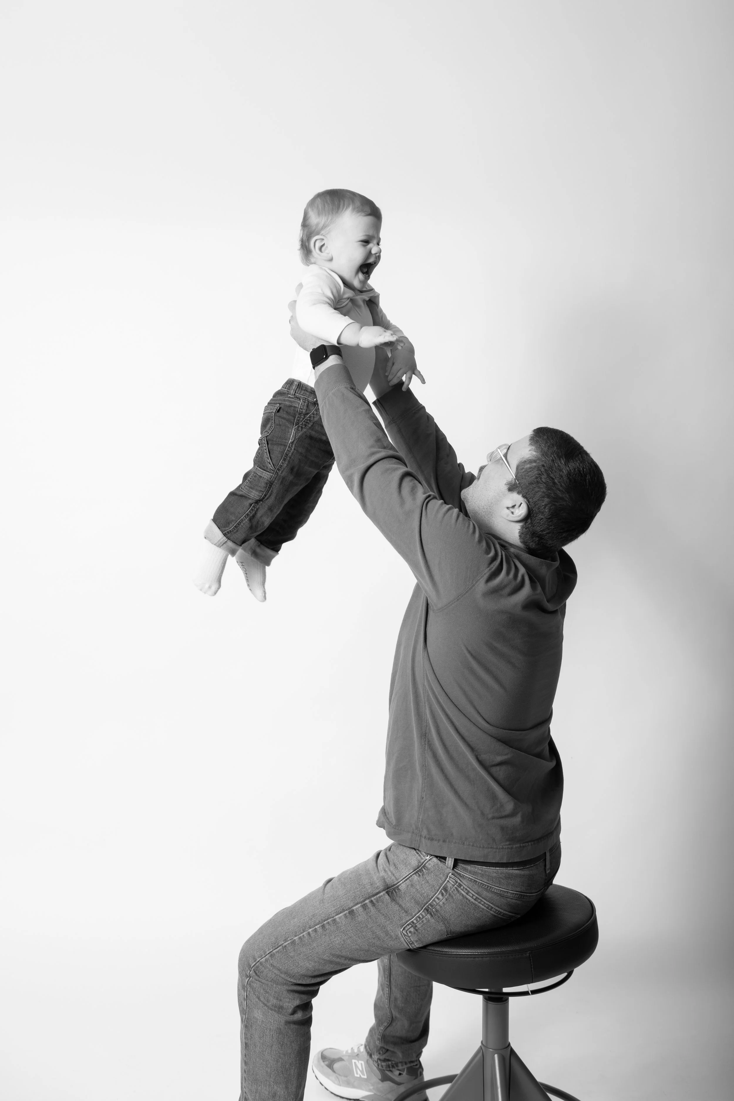 A man sitting on a stool lifting a laughing baby in the air against a plain background.