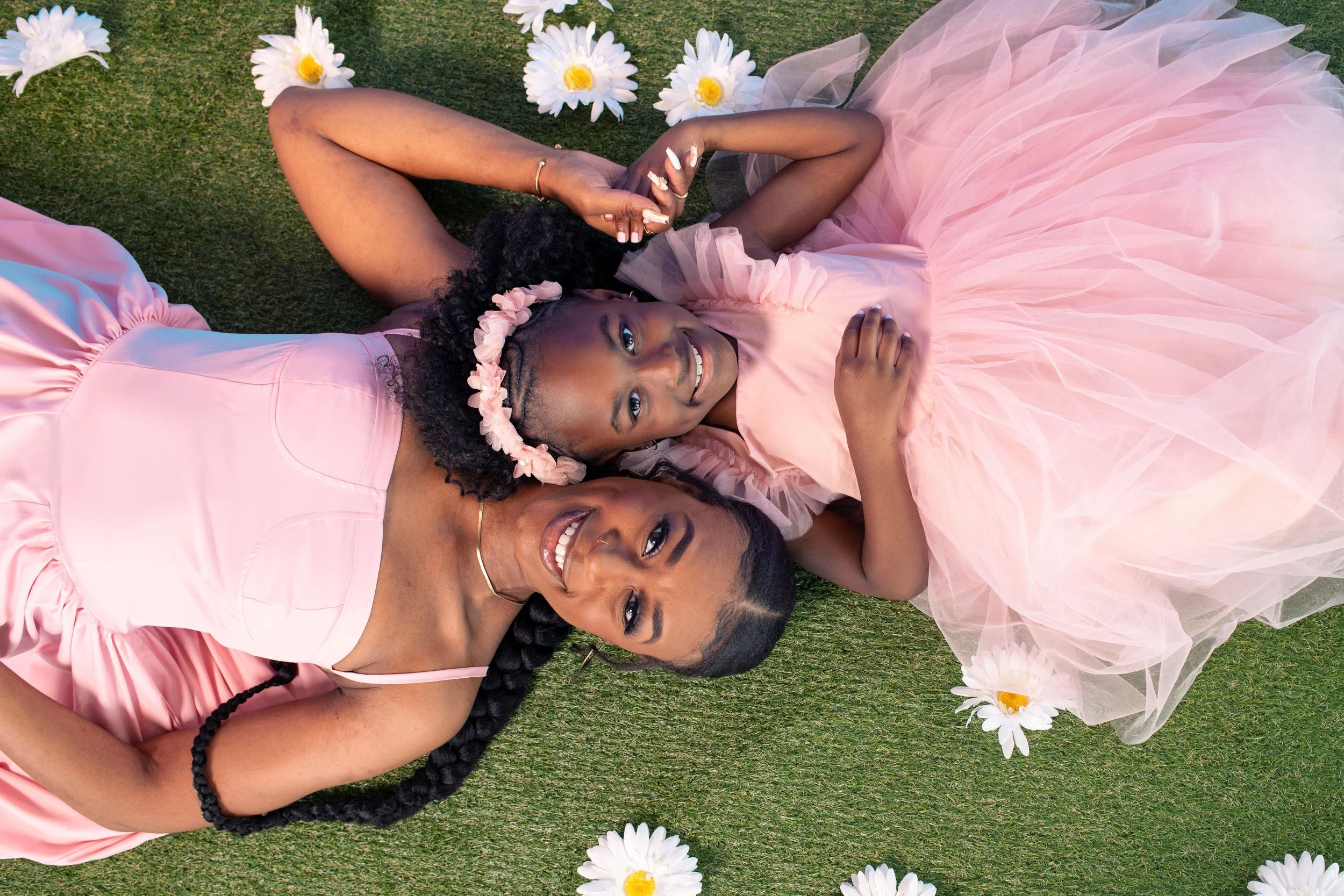 Two women lying on grass, one in a pink dress with braid and one in a pink tulle dress with flower crown, surrounded by white daisies.