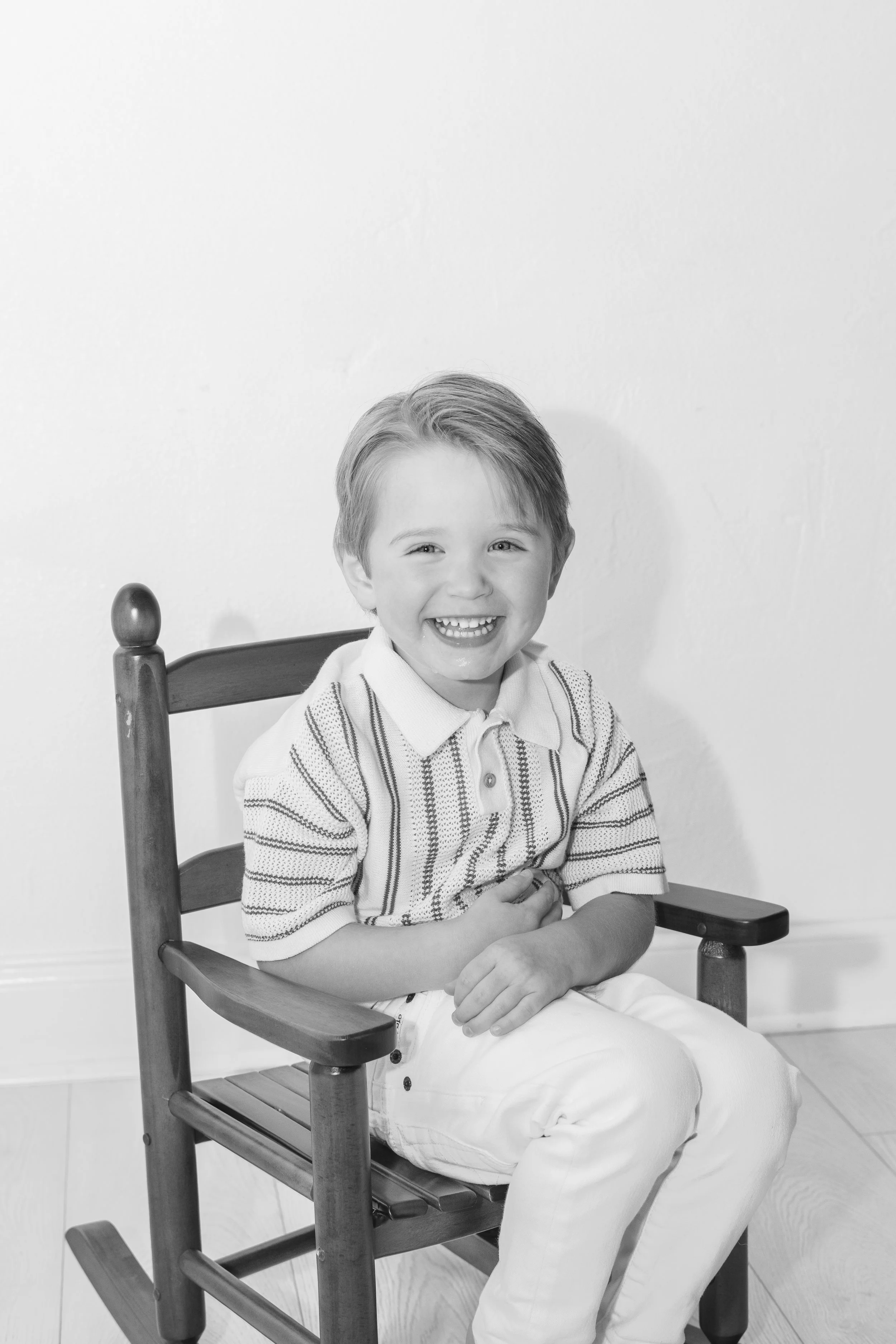 A smiling young boy sitting on a wooden chair against a plain wall, wearing a striped polo shirt and light-colored pants.