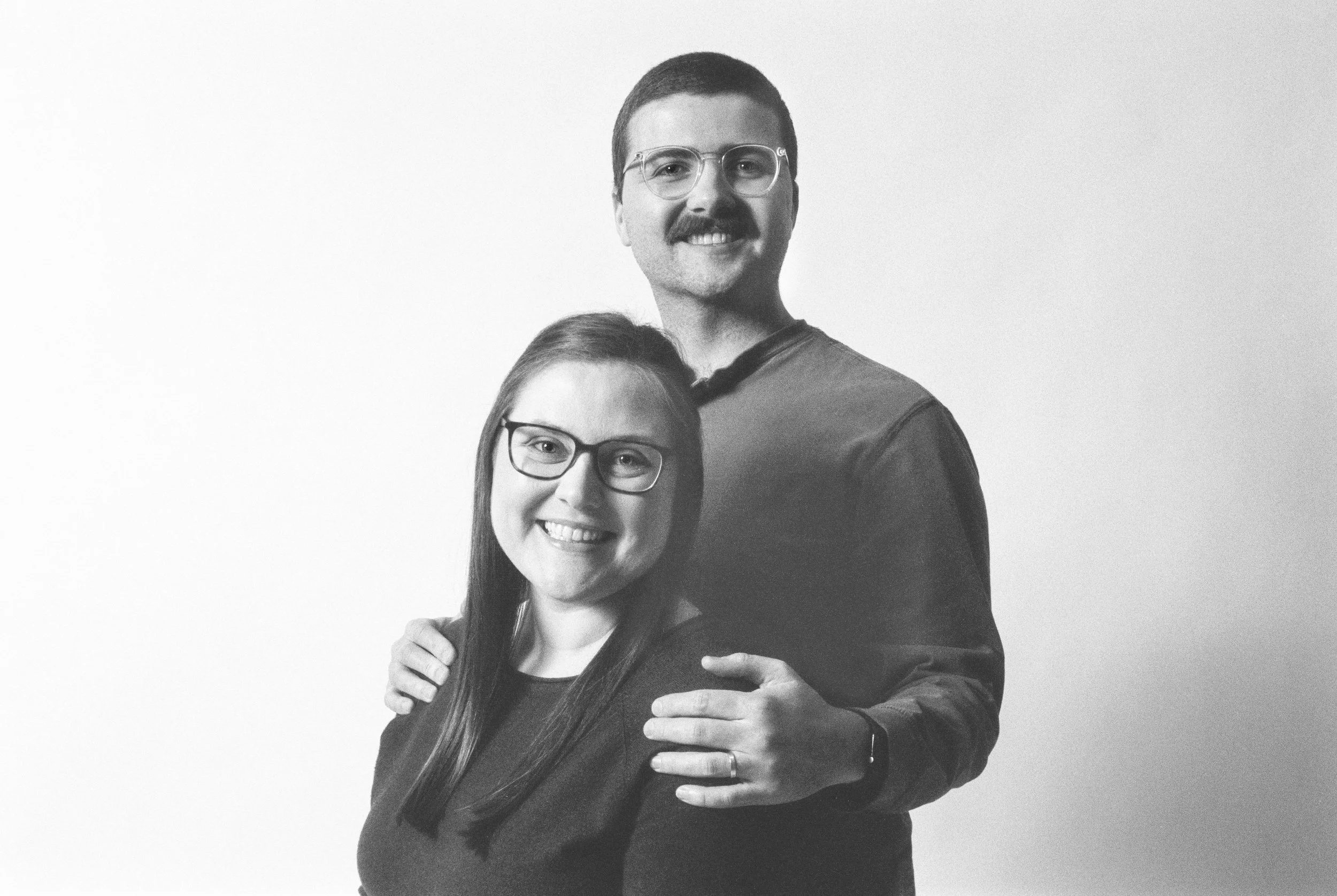 A smiling couple with glasses, standing close together, with the man's hand on the woman's shoulder, against a plain light background.