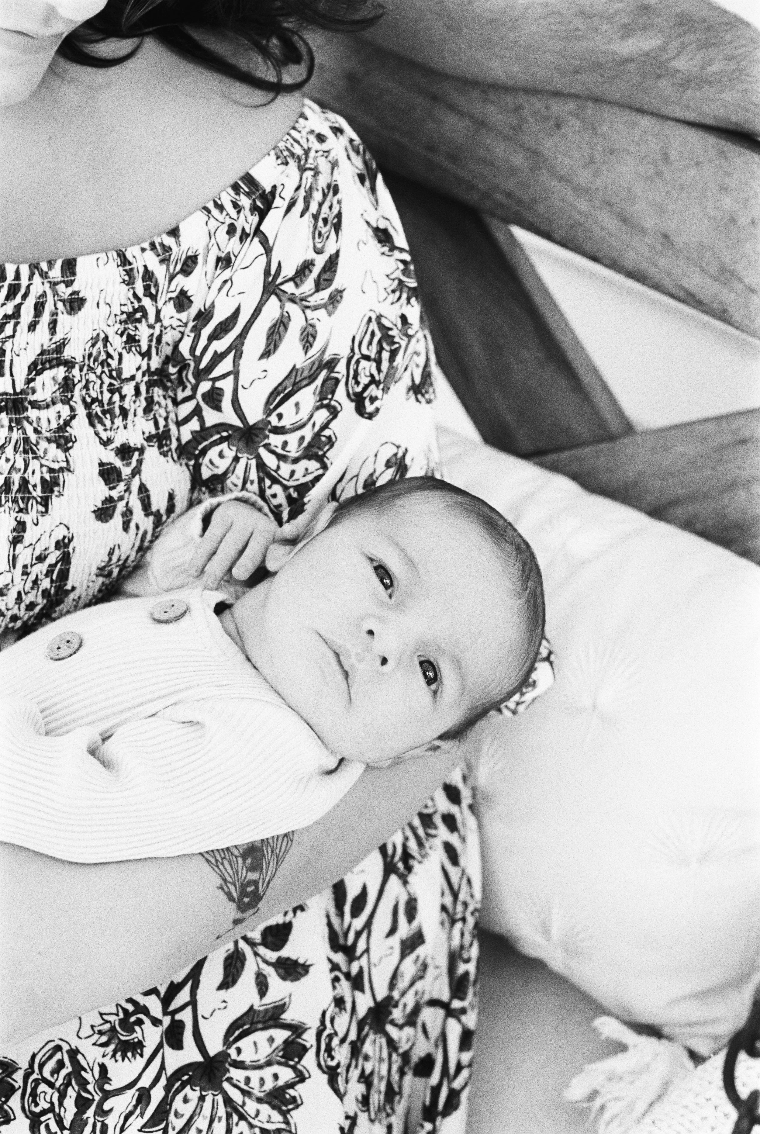A baby lying on a person's chest, looking at the camera, with a patterned fabric and a pillow in the background.