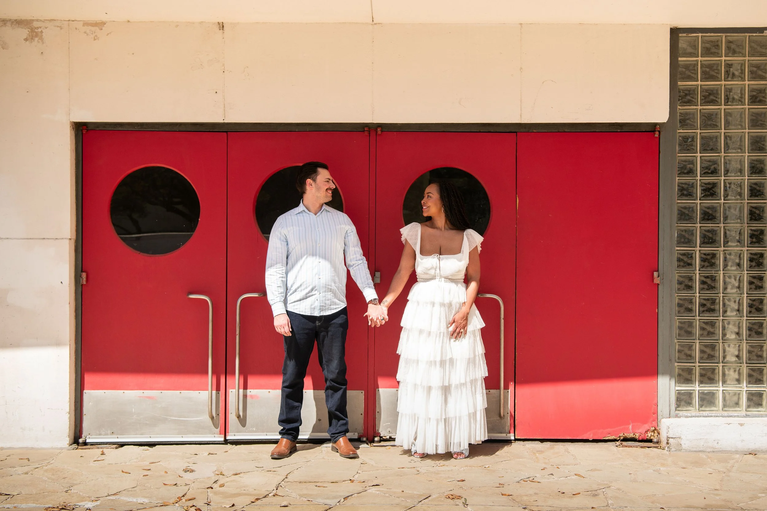 A couple holding hands and smiling at each other standing in front of a red door with circular windows.