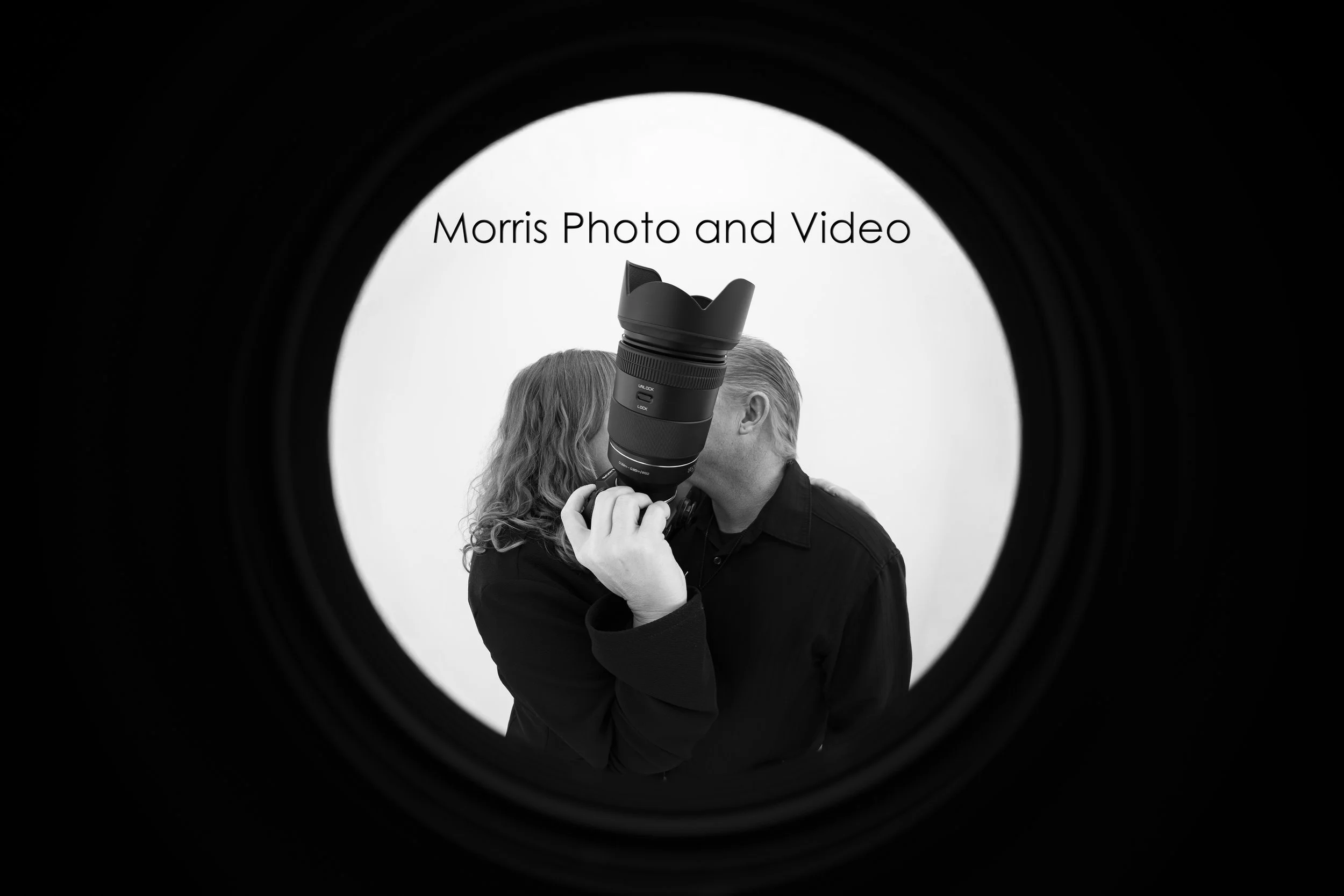 Black and white image of a woman and a man seen through a camera lens, with the woman holding a camera lens to the man's eye, creating a visual effect that frames the scene.