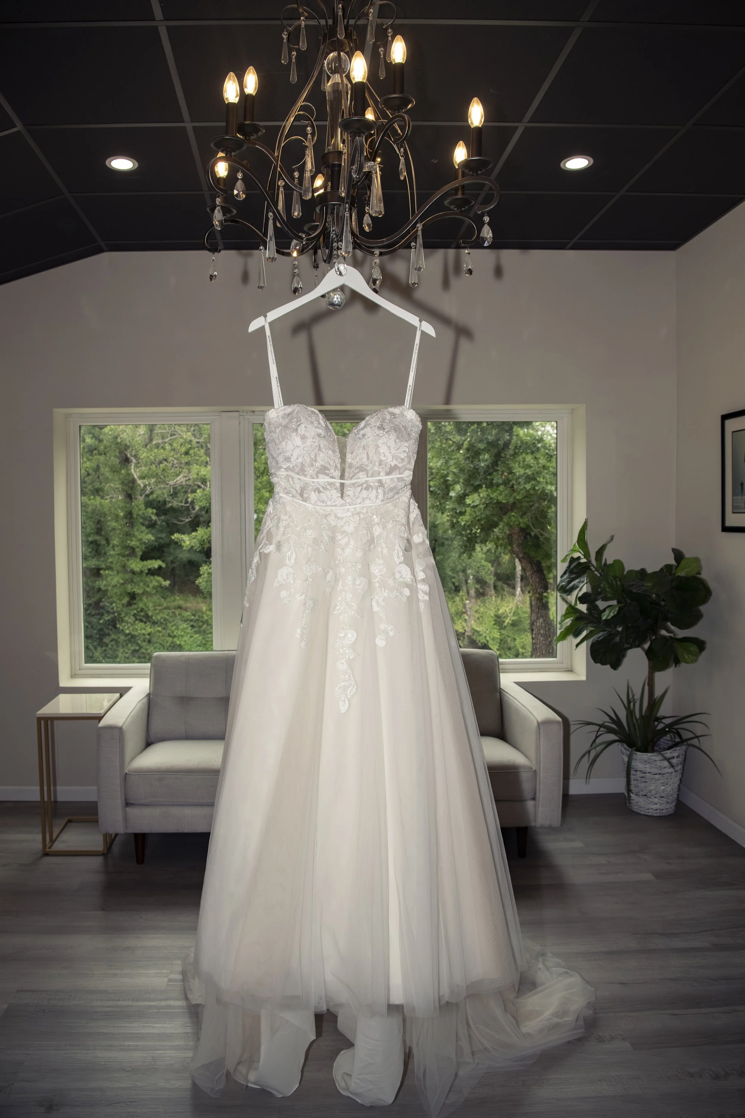 A wedding dress hanging from a chandelier in a room with a large window and views of green trees outside.