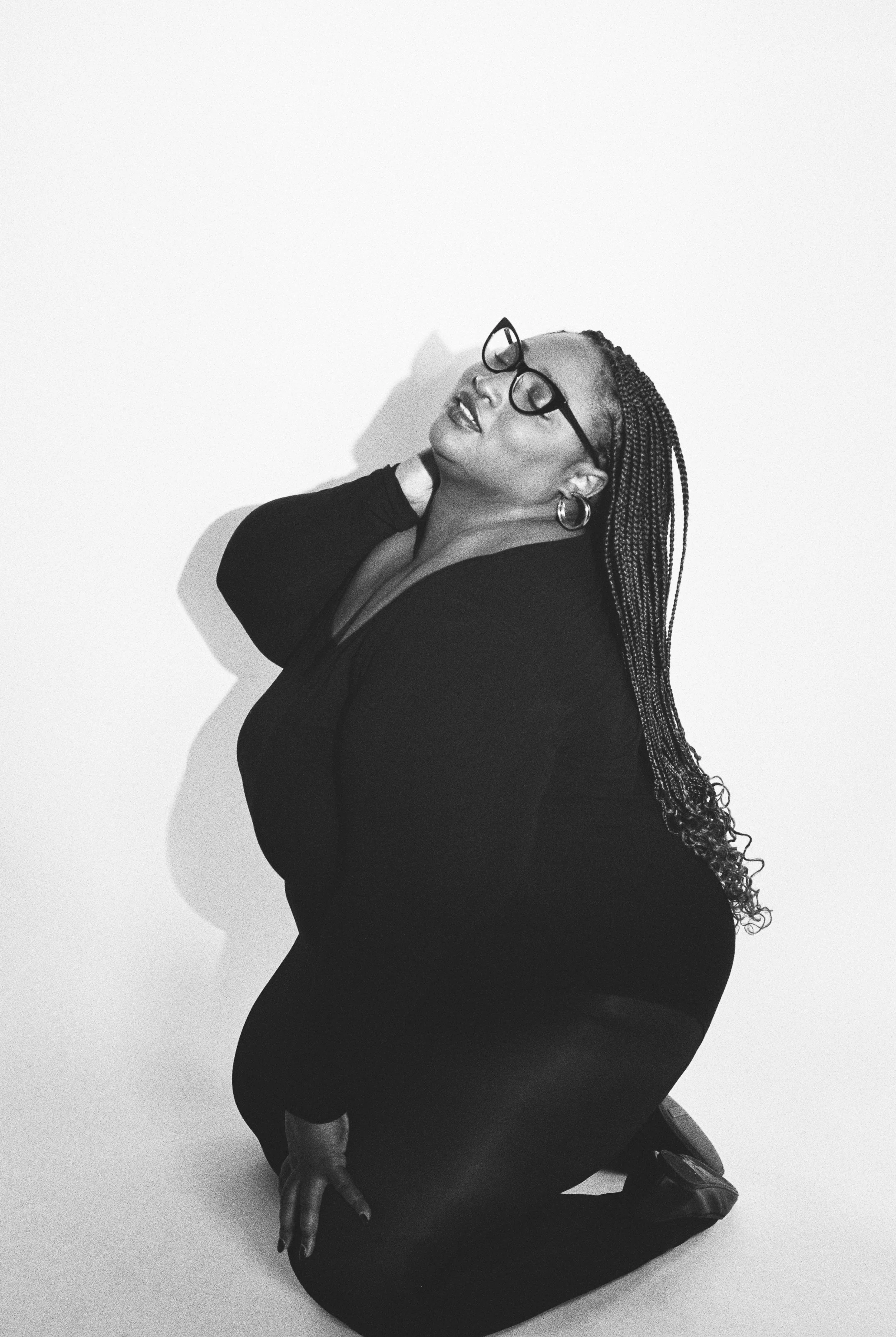 Black and white photo of a woman kneeling on the floor with one hand resting on her neck, wearing glasses, hoop earrings, and a dark outfit, with her head tilted back and eyes closed.
