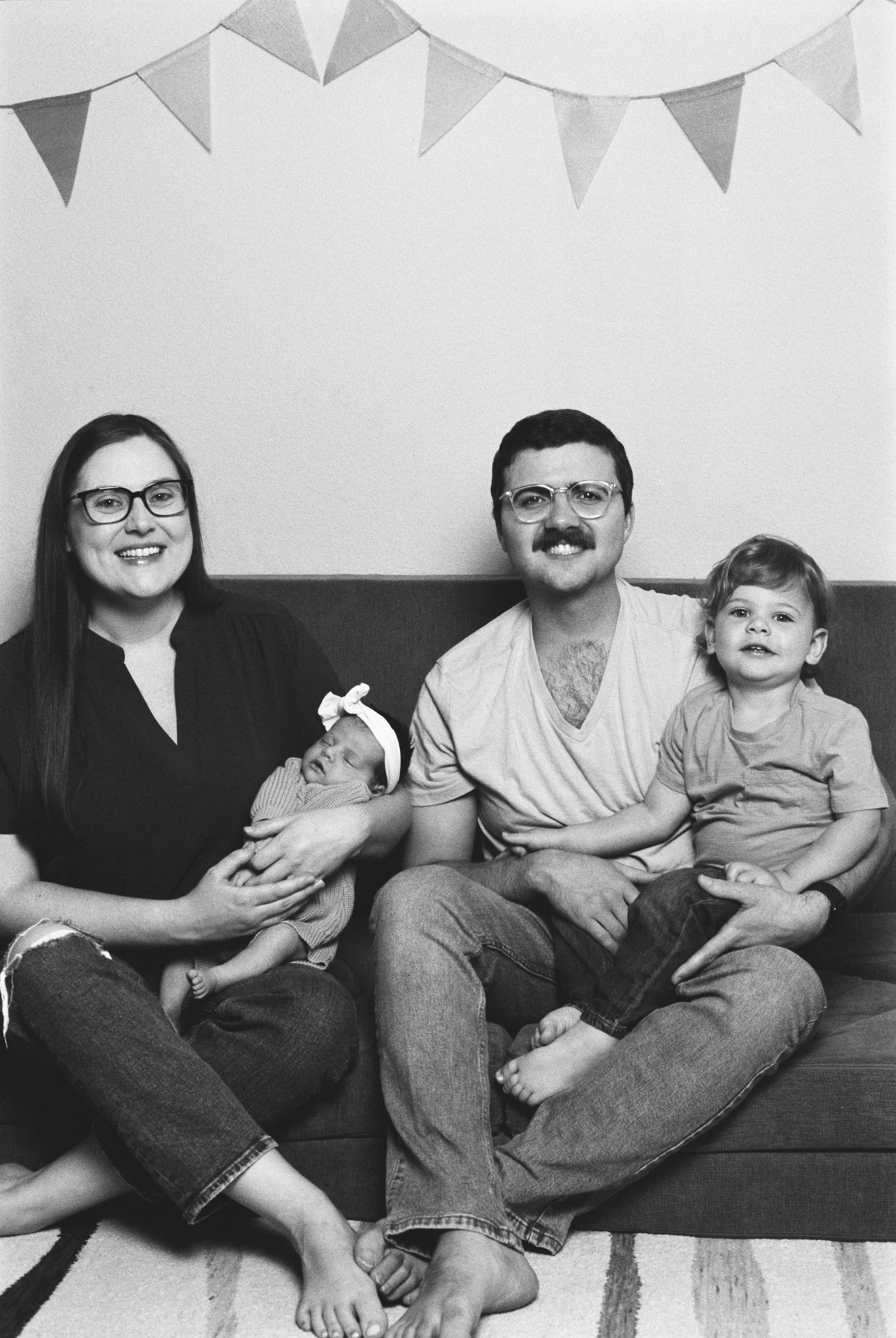 A family portrait of four sitting on a couch, including a woman holding a newborn baby, a man, and a young boy, with bunting decorations hanging above them.