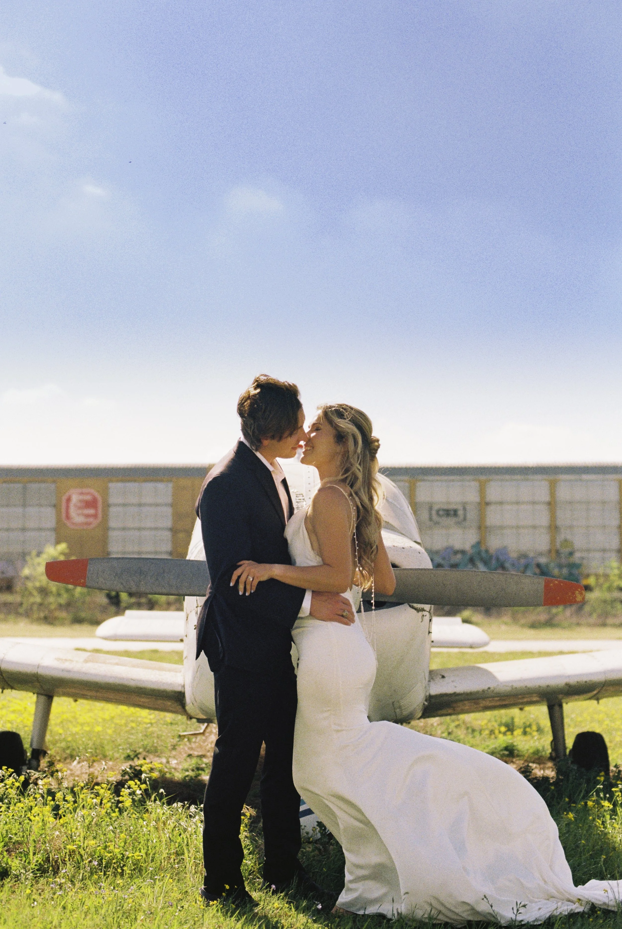 A bride and groom sharing a kiss in front of an airplane, with the bride kneeling and the groom standing, holding the bride's waist.