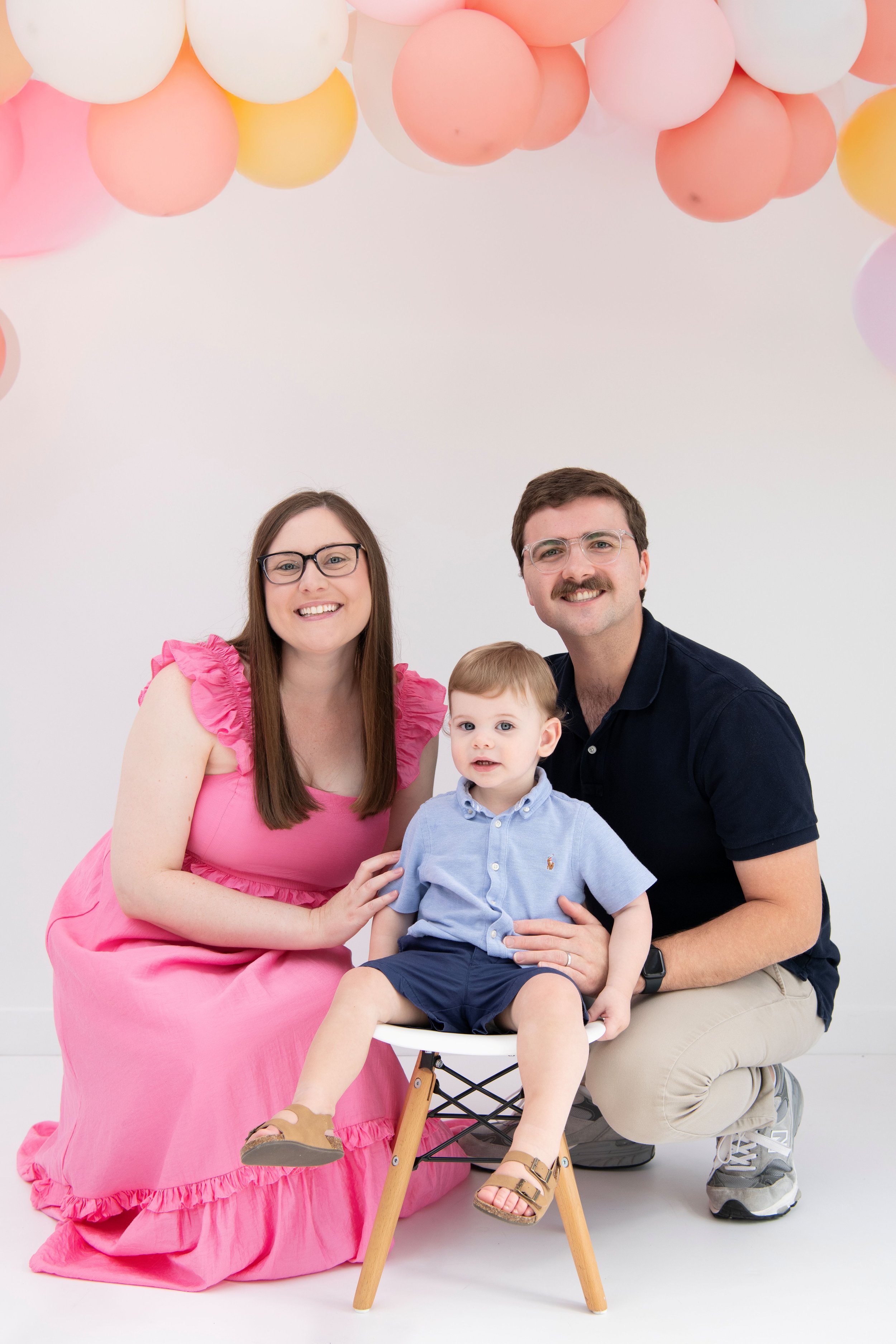 A family of three, a woman, a man, and a young boy, smiling and posing for a photo in front of a white background with pink, white, yellow, and purple balloons hanging from the top.