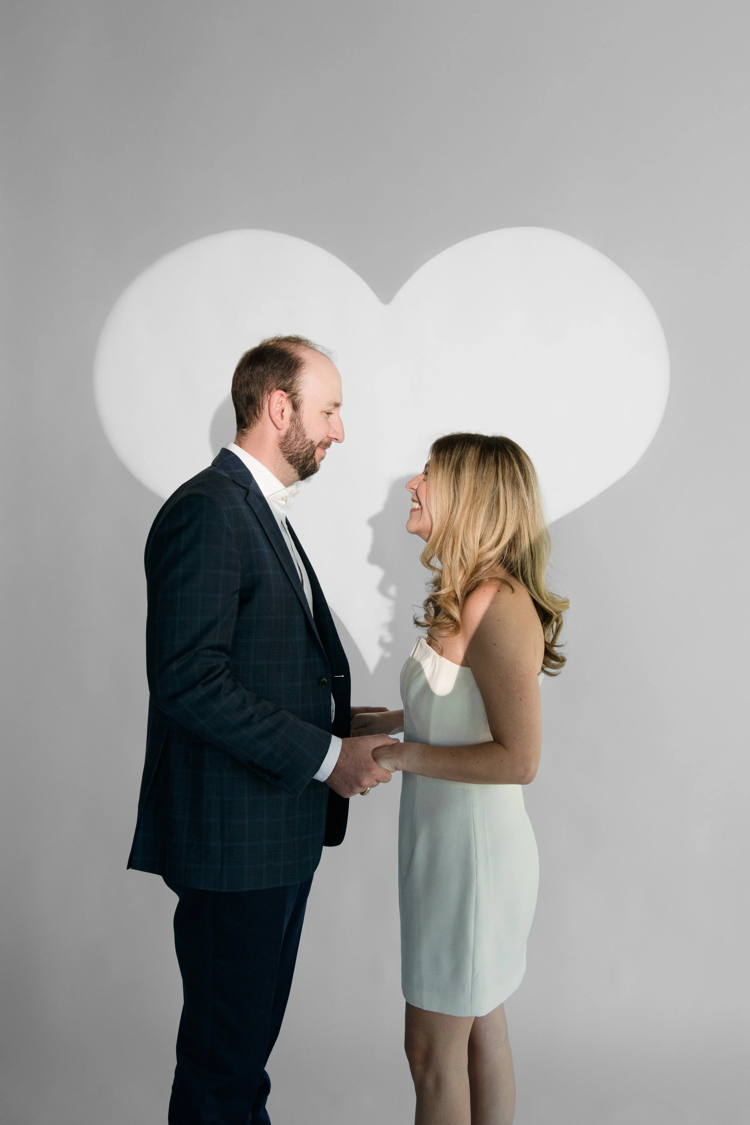 A couple holding hands and smiling at each other, standing in front of a background with a large white heart projected on the wall.