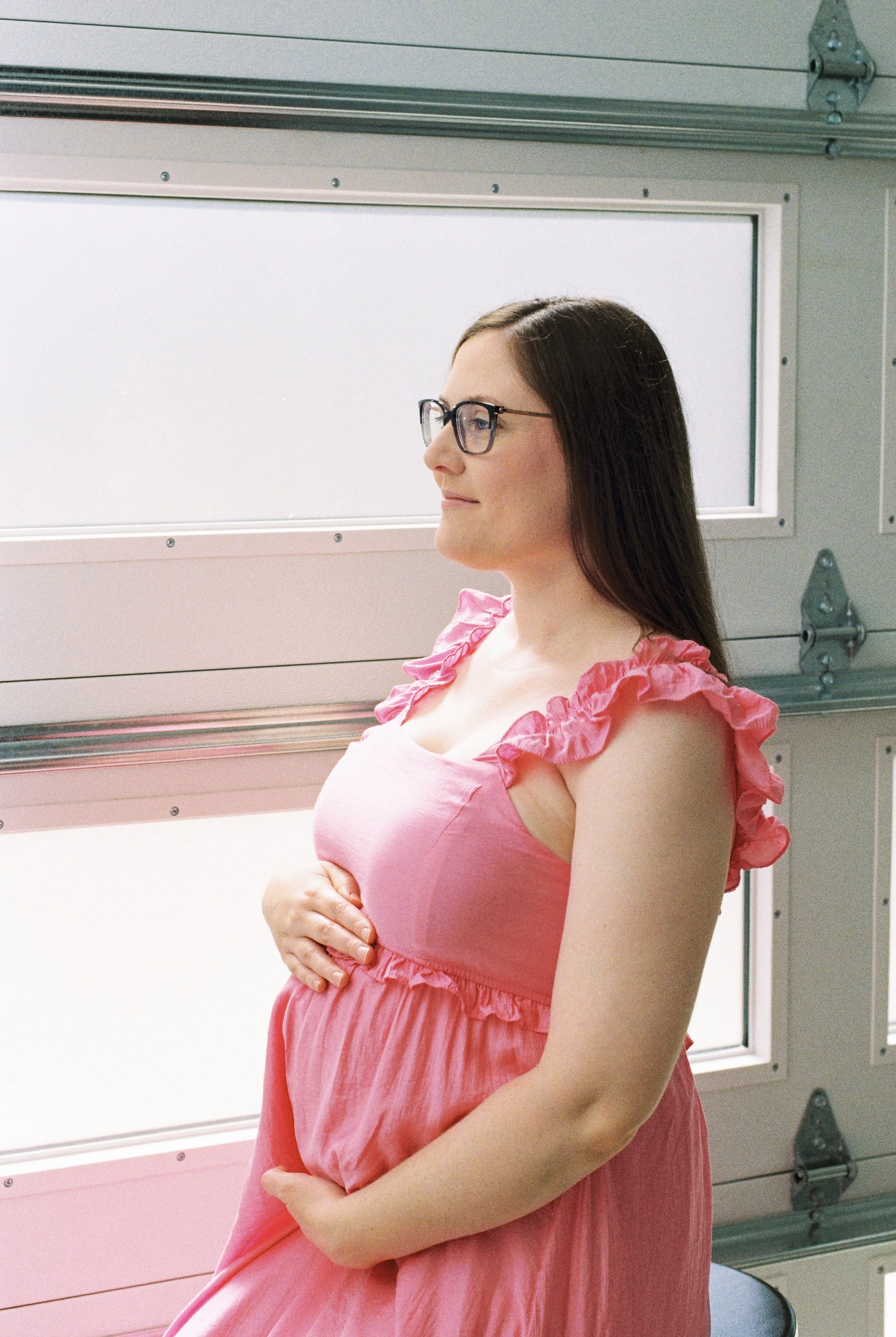 A pregnant woman with glasses, wearing a pink dress with ruffled shoulders, sitting and holding her belly in a room with pink and gray panels.