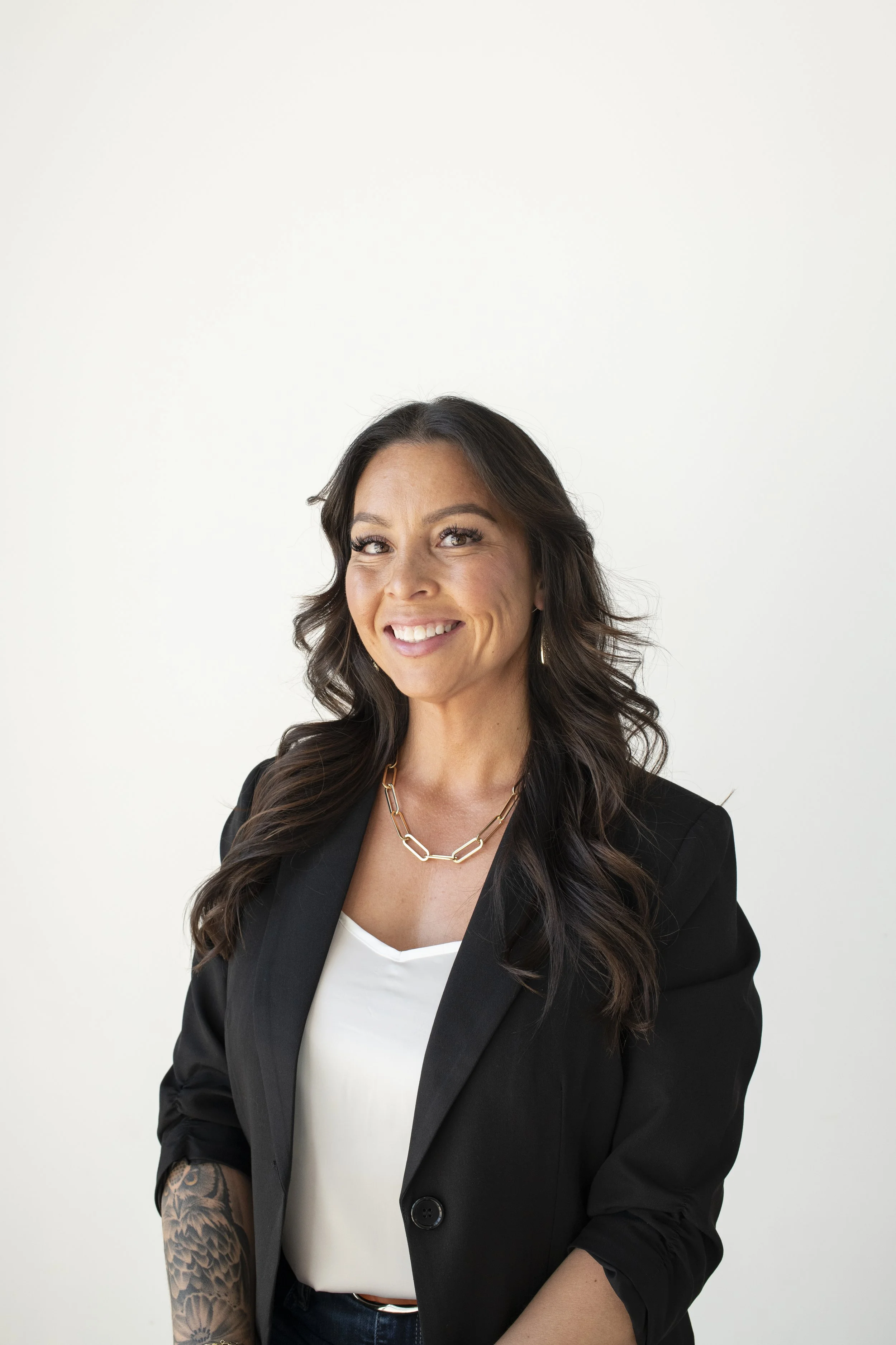 A woman with long wavy dark hair, wearing a black blazer, white top, and a gold chain necklace, smiling in front of a plain white background.