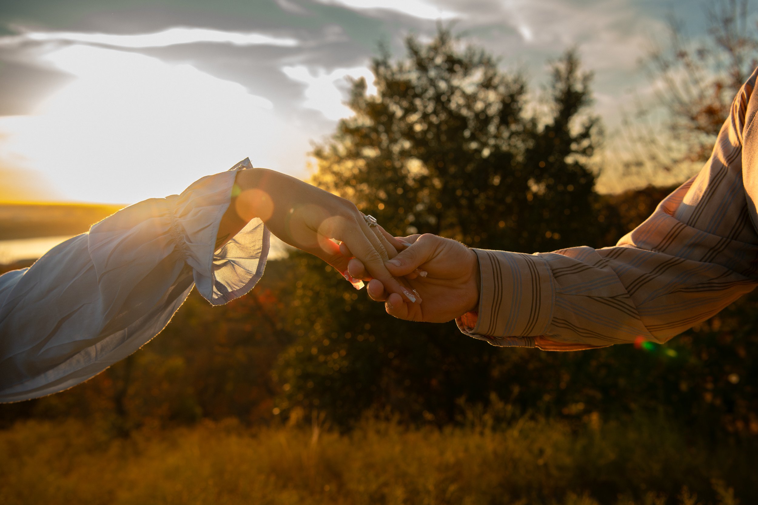 Two people holding hands outdoors during sunset, with trees and a bright sky in the background.