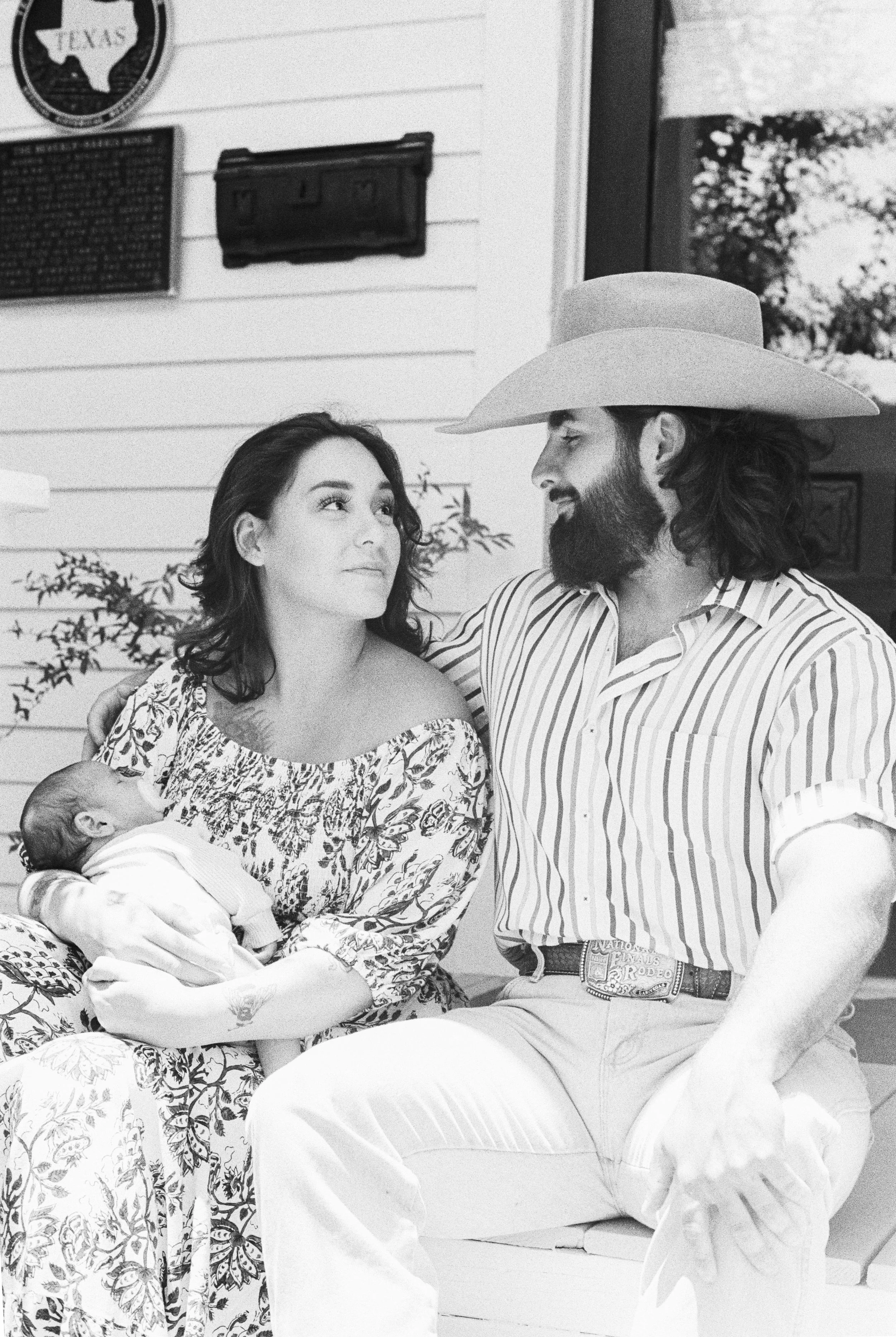 A woman holds a baby in her arms while a man in a cowboy hat and striped shirt sits beside her, looking at her lovingly. They are seated outdoors, with parts of a white wooden house and a sign visible in the background.