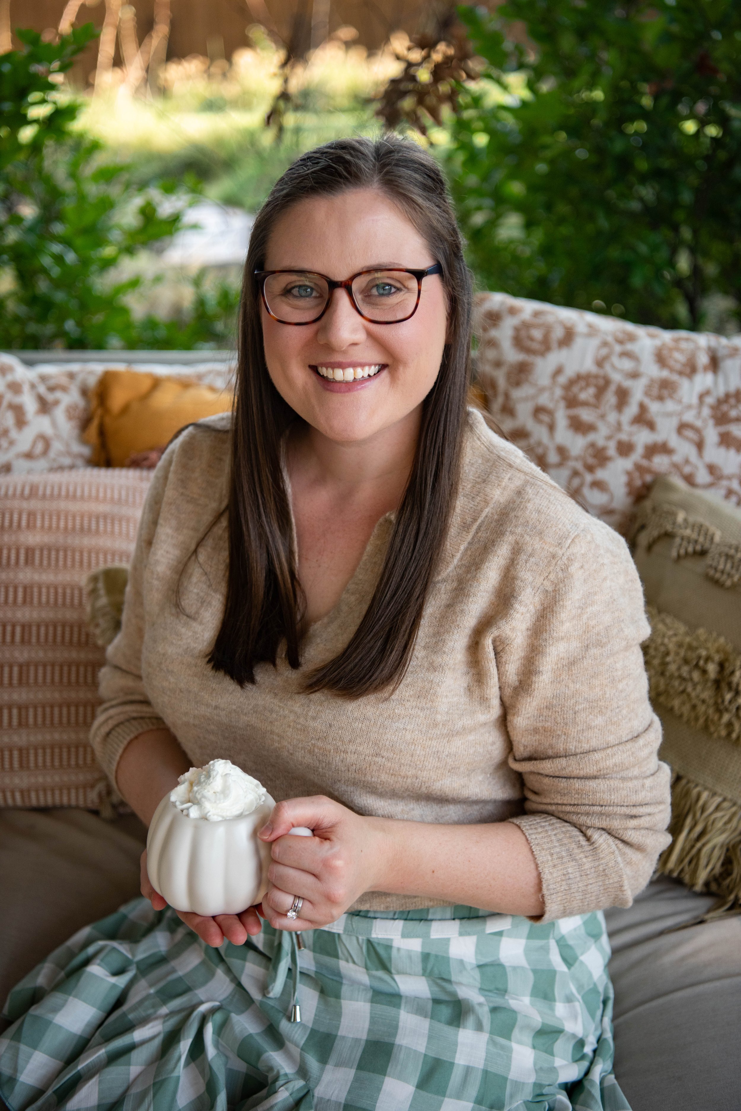 A woman with long brown hair, glasses, and a warm smile, sitting indoors with a background of patterned pillows and greenery outside. She is holding a cup filled with whipped cream.