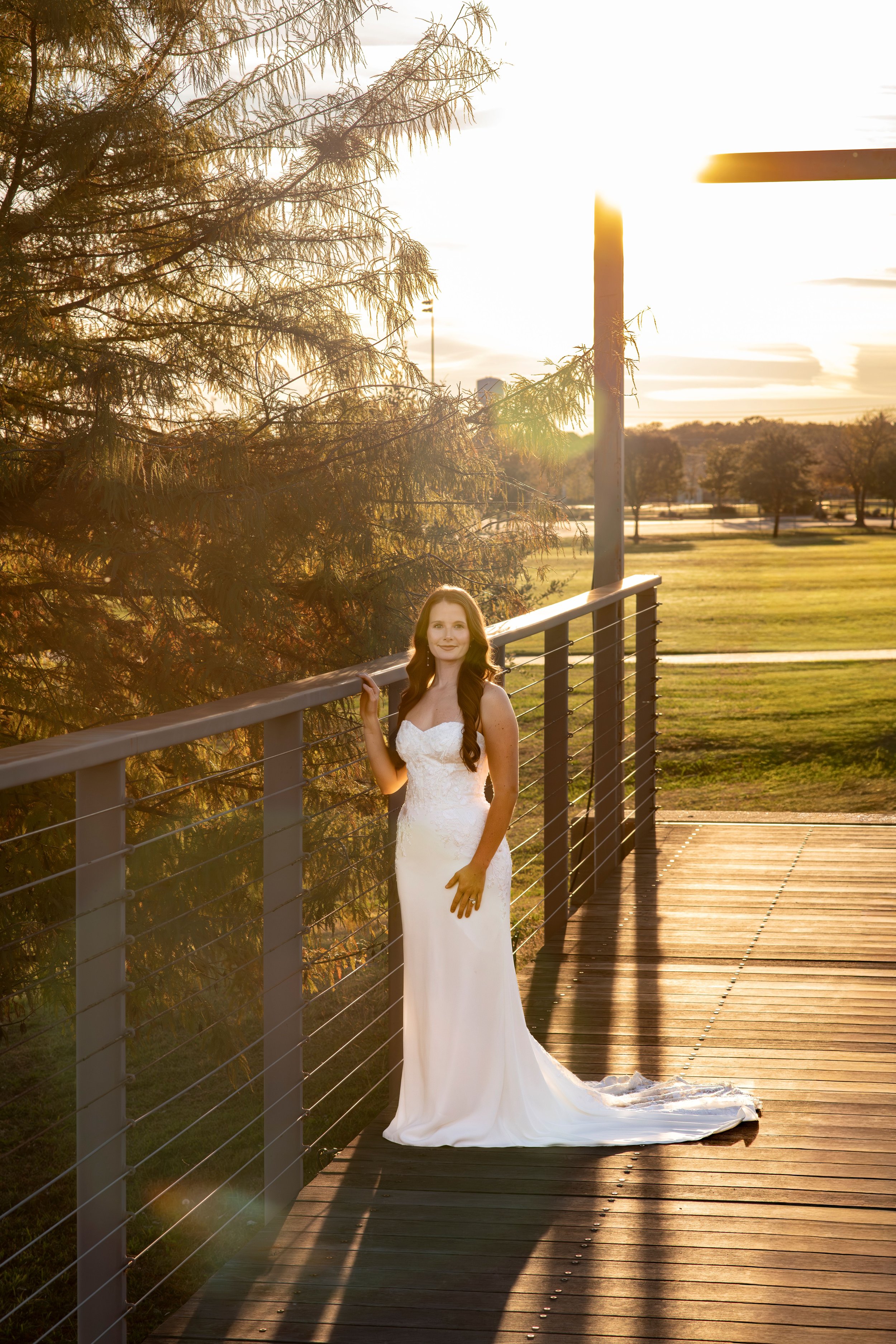 A woman in a white wedding dress standing on a wooden bridge at sunset.